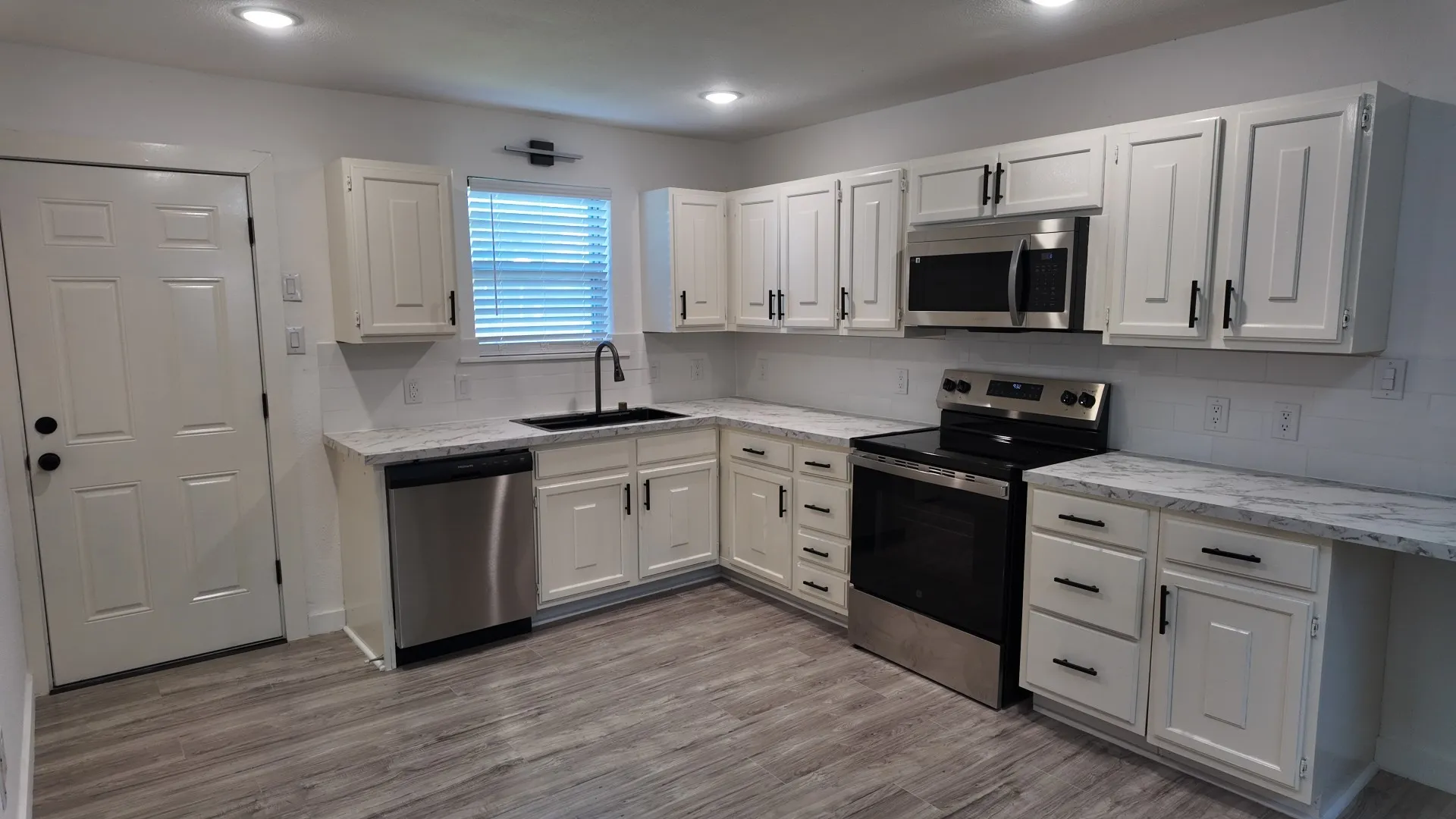 Kitchen featuring white shaker-style cabinetry, stainless steel appliances, a black sink with a matte black faucet, light-toned countertops, and wood-finish flooring