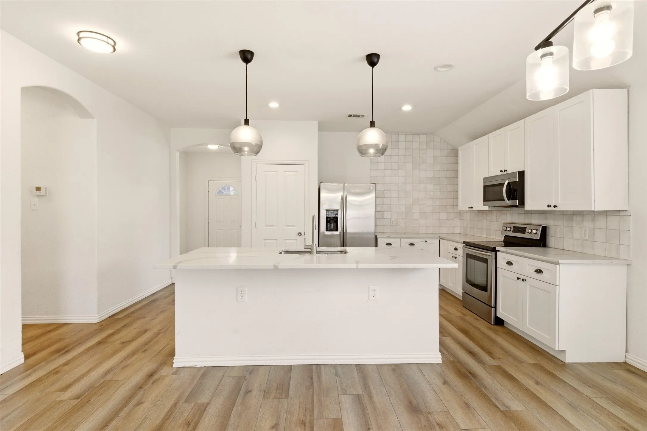 Kitchen featuring an island with a built-in sink, stainless steel appliances, white cabinetry, tile backsplash, and wood-finish flooring