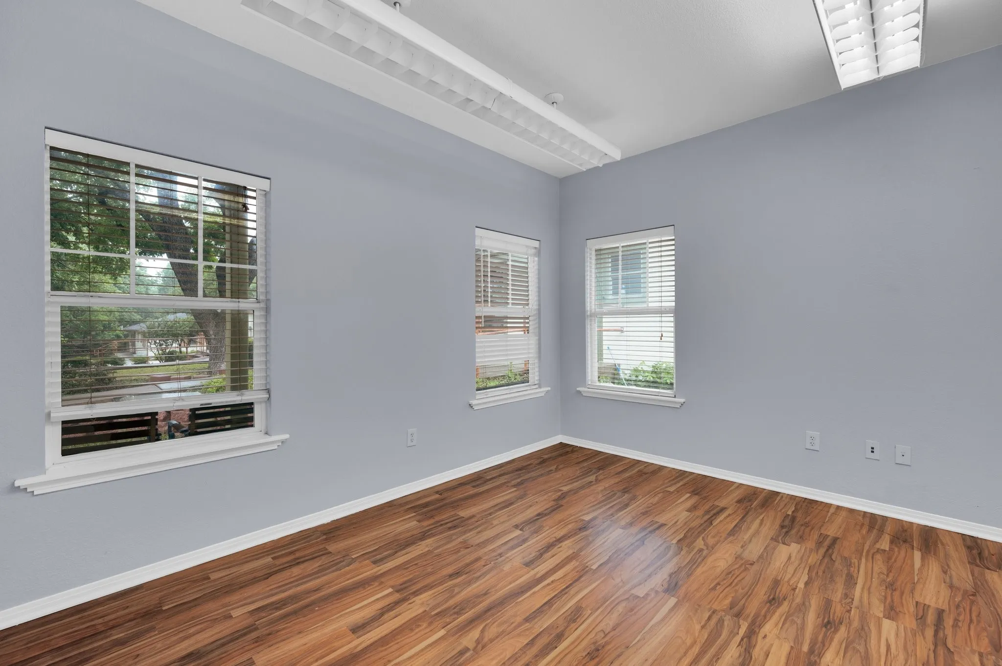 Wood-finish flooring with white trim, multiple windows with white blinds, and overhead lighting fixtures