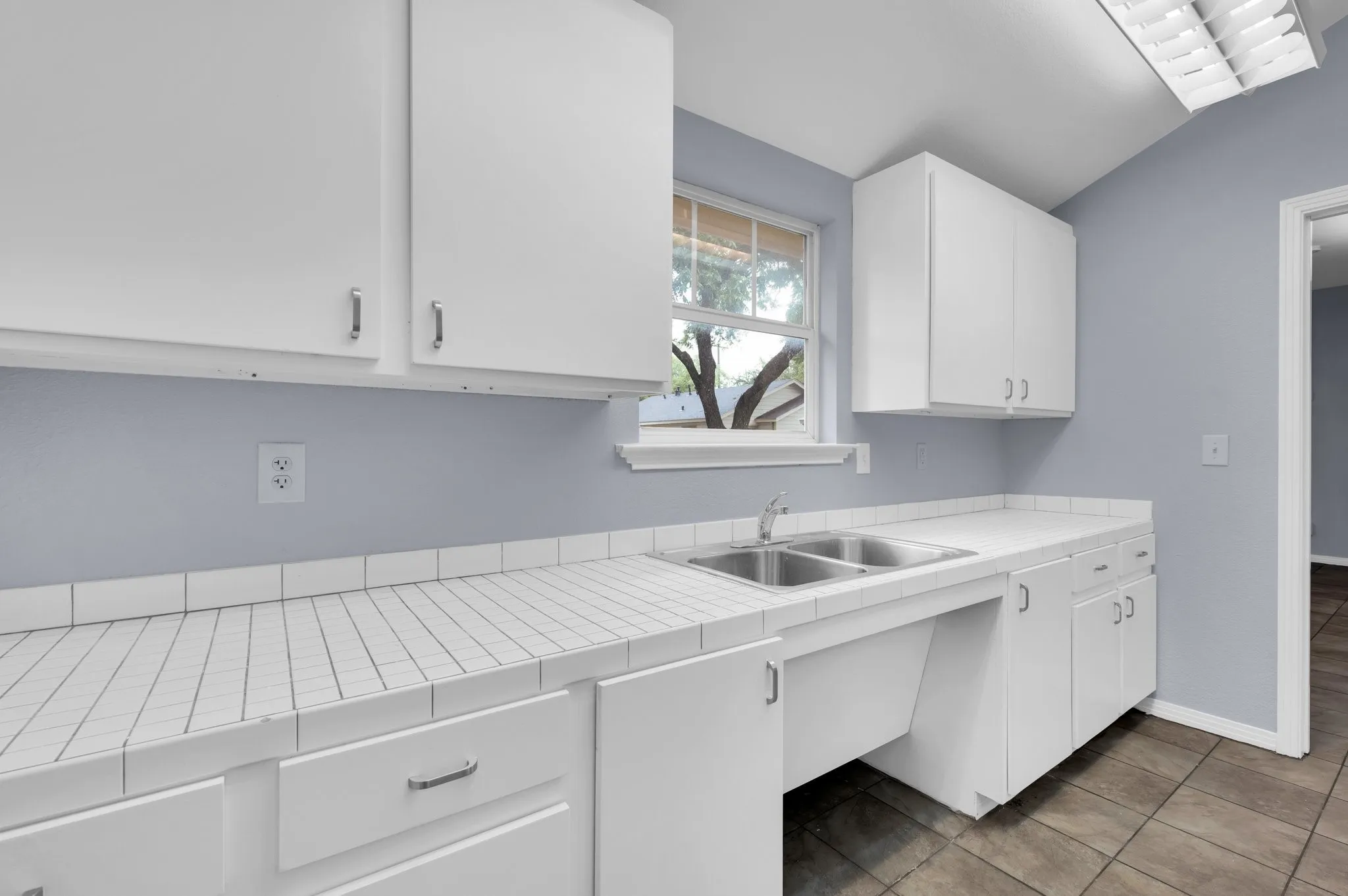 White cabinetry, tile countertops, stainless steel double-basin sink, and a window with exterior visibility