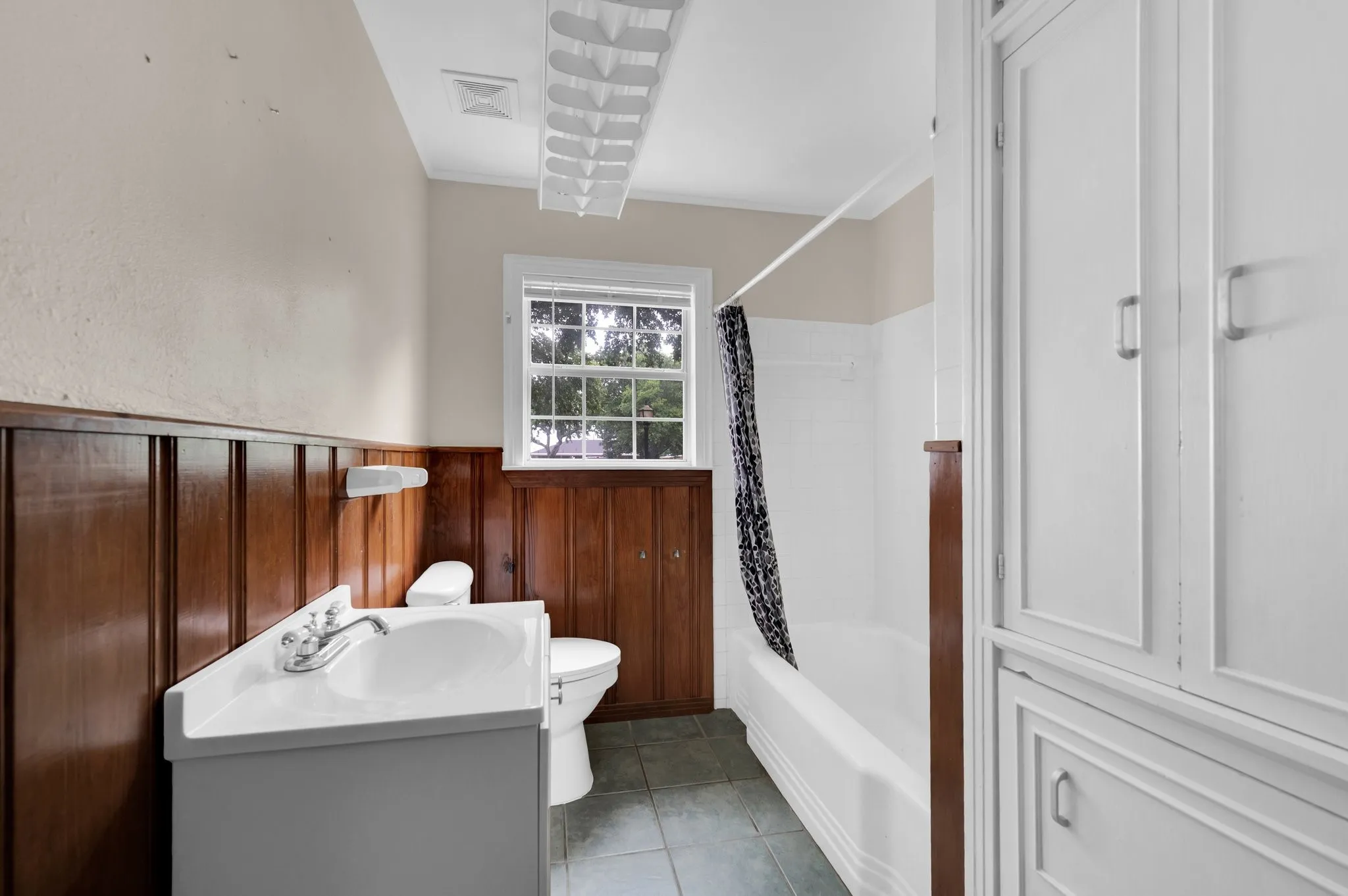 White vanity with integrated sink, wood-paneled wainscoting, white bathtub, tiled flooring, and built-in white cabinetry