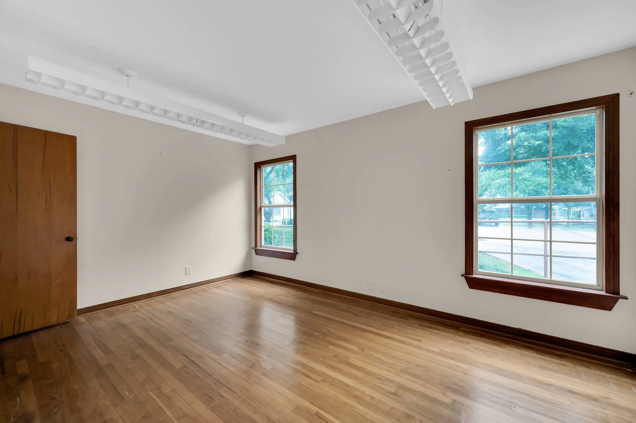 Hardwood flooring, two large windows with wood trim, ceiling-mounted fluorescent lighting, and a wood-panel door