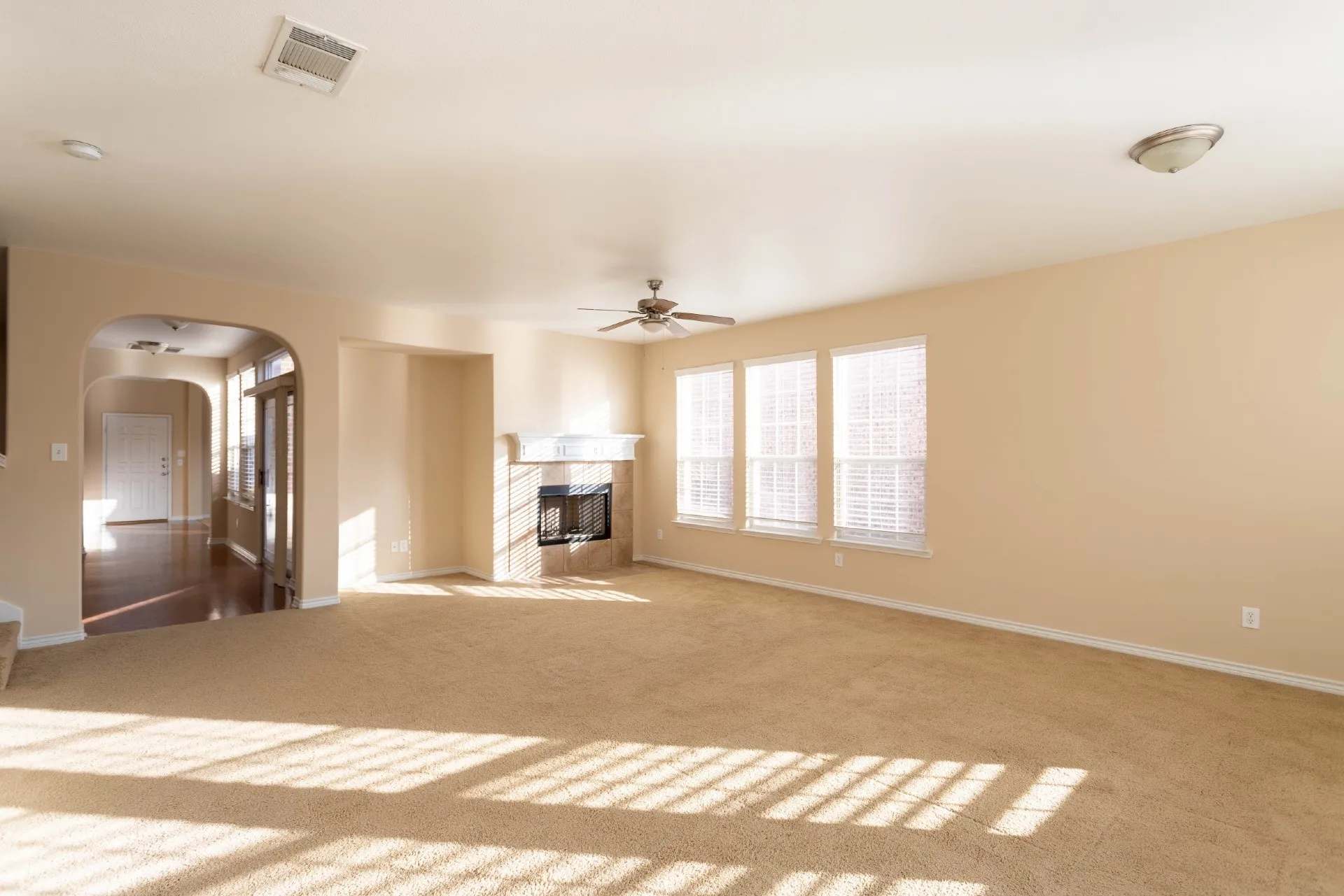 Carpeted living area featuring a fireplace with a stone surround, three windows with blinds, a ceiling fan, and an arched entry