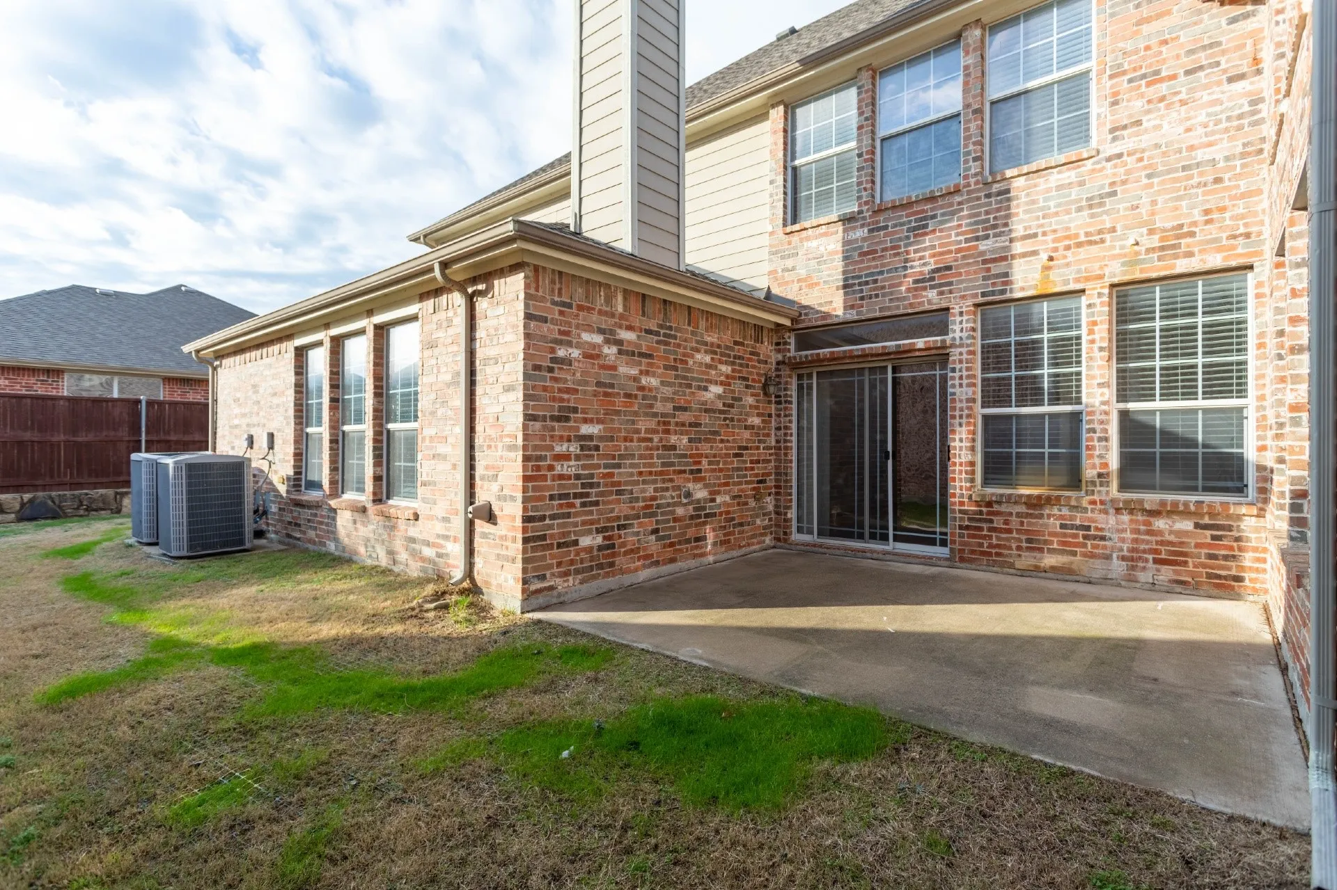 Brick exterior with multiple windows, concrete patio, sliding glass door, and a grassed yard area