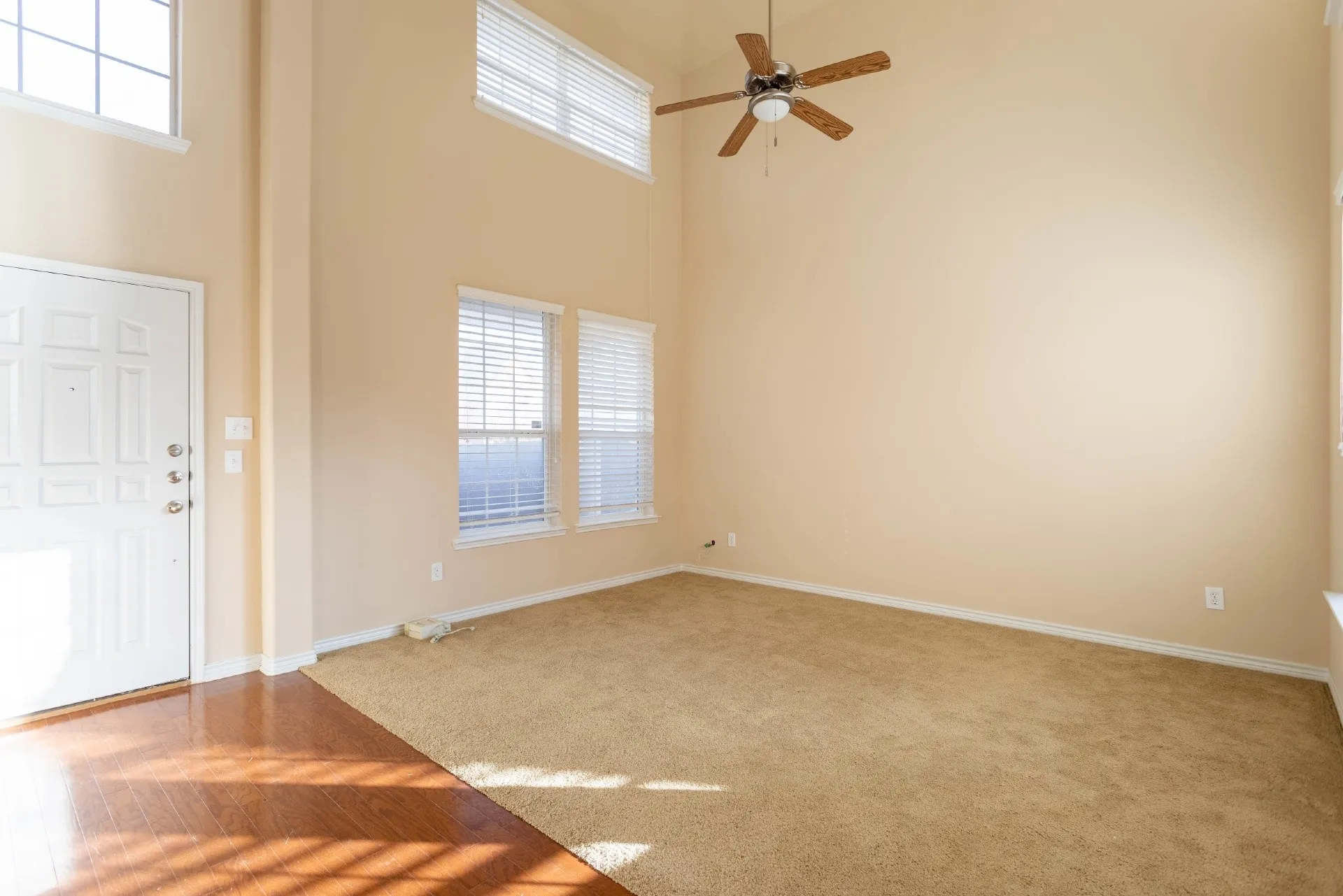 Two-story ceiling, multiple windows with blinds, ceiling fan, wood-finish flooring, and light-colored carpet