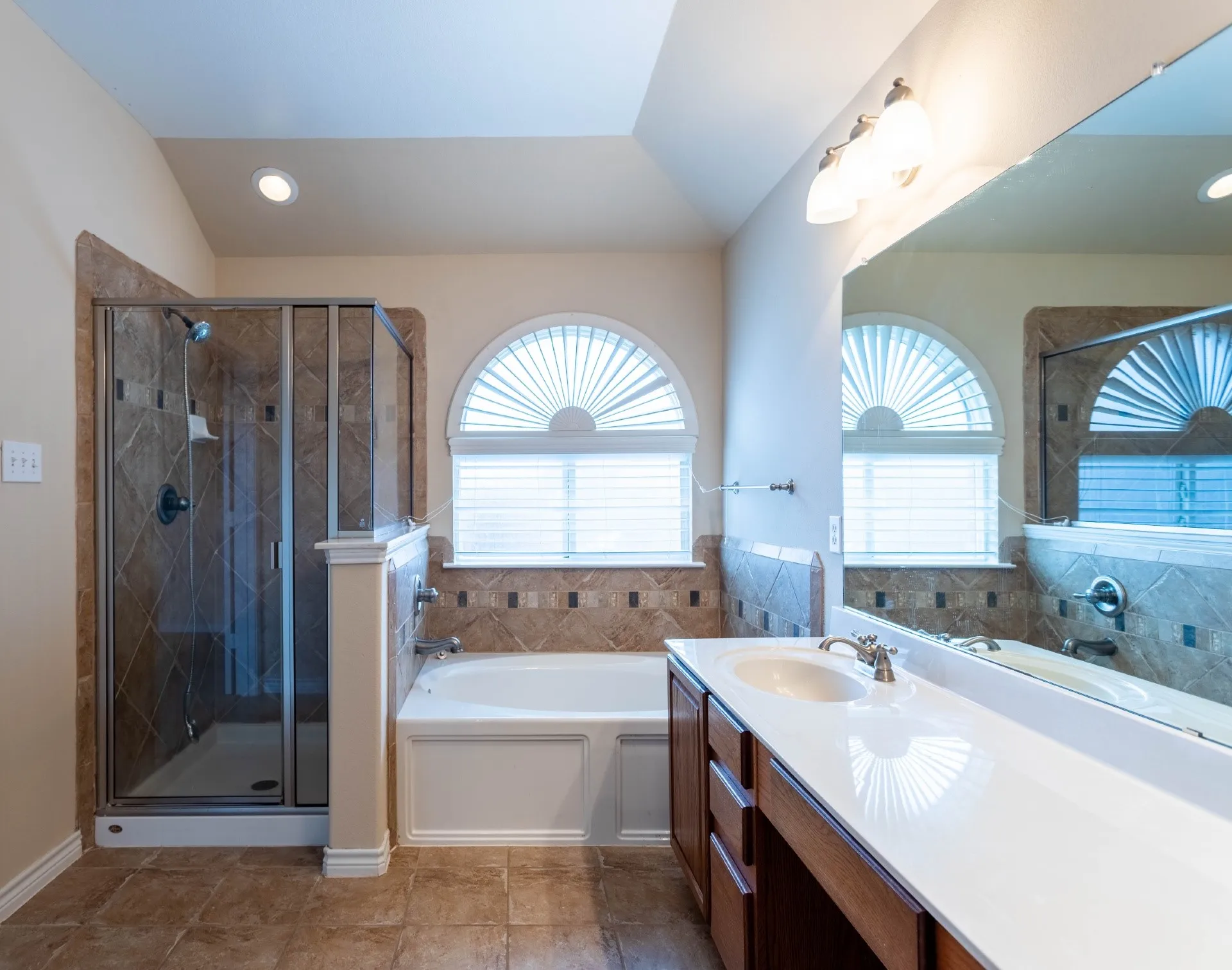 White bathtub with arched window, glass-enclosed shower, wood-finish vanity with white countertop, and tiled flooring