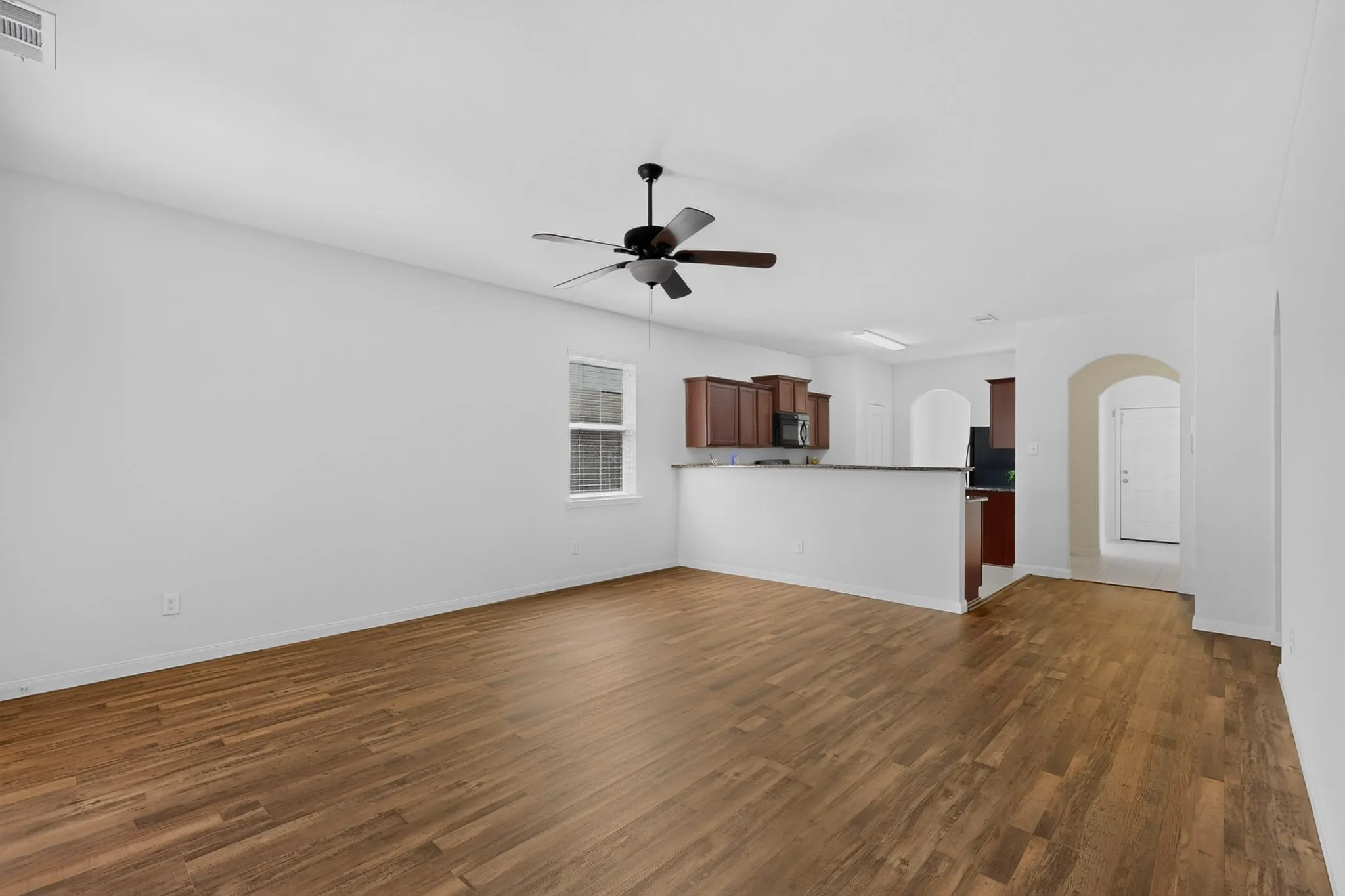 Spacious living area with wood-finish flooring, a ceiling fan, and a pass-through to the kitchen featuring dark wood cabinetry and a breakfast bar
