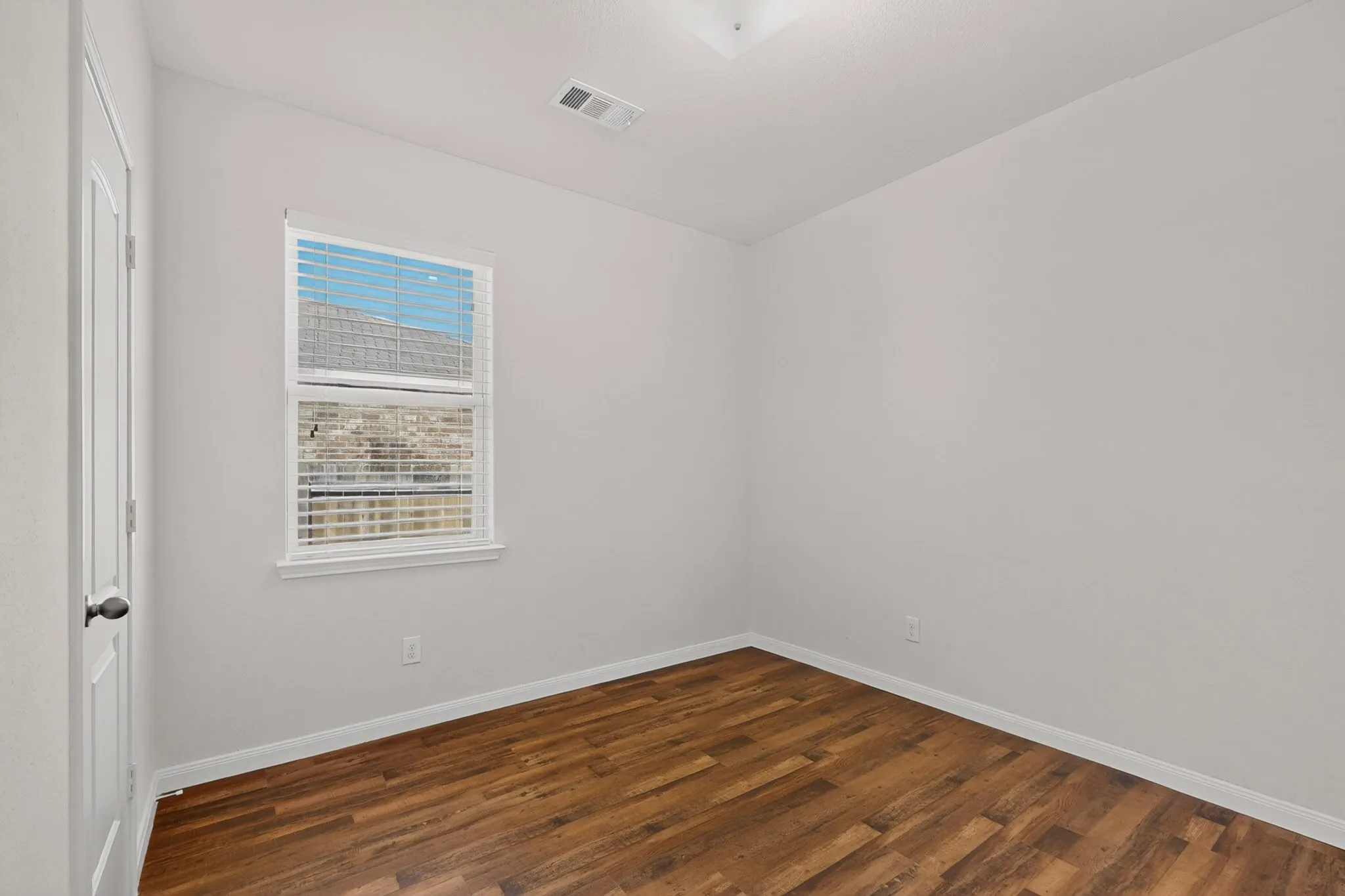 Wood-finish flooring with white baseboards, a single window with blinds, and a white interior door with a black knob