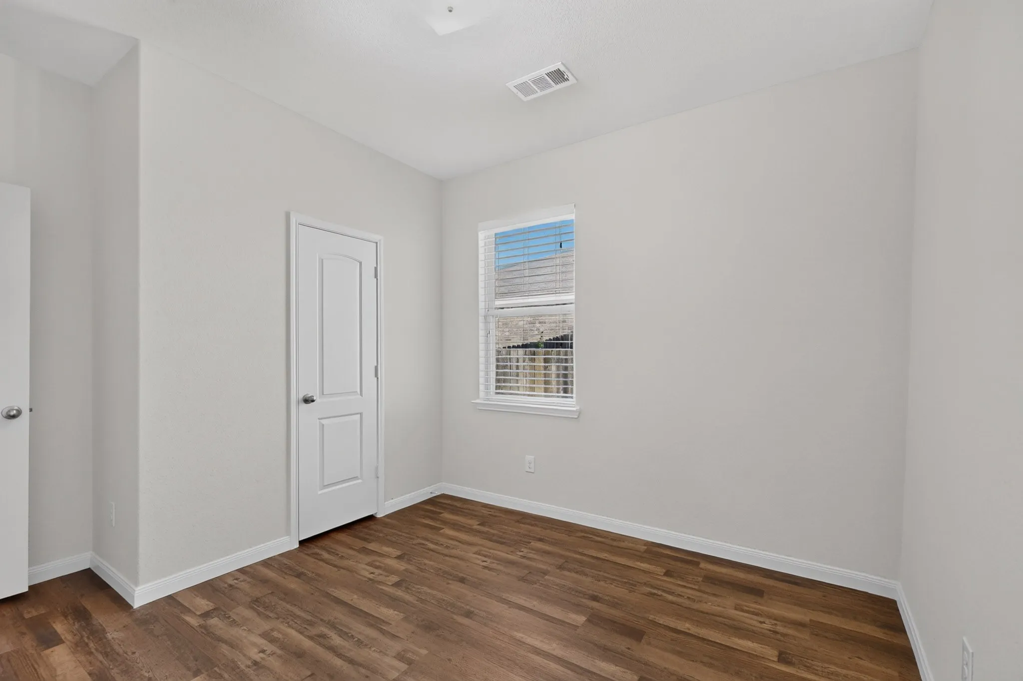 Wood-finish flooring, white paneled door, white trim, single window with blinds, and light-toned walls