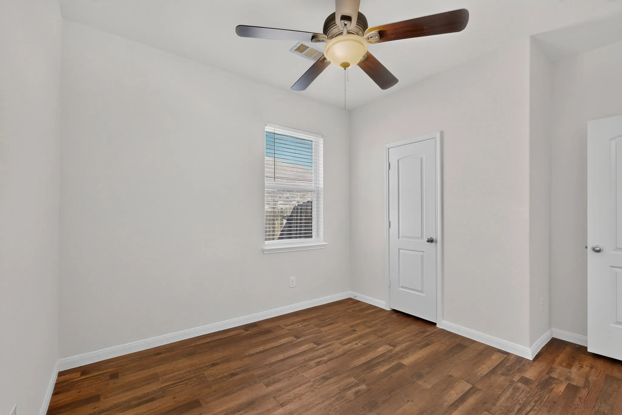 Light-filled room featuring wood-finish flooring, a white window with blinds, a ceiling fan with light fixture, and two white panel doors
