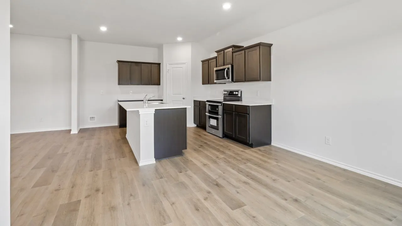 Open-concept kitchen featuring an island with an integrated sink, dark wood cabinetry, stainless steel appliances, a white tile backsplash, and light wood-finish flooring