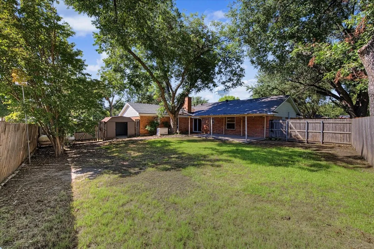 Expansive backyard featuring a covered patio, brick exterior, wood privacy fencing, mature trees, and a utility shed