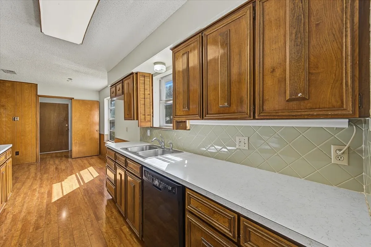 Wood-finish flooring, wood cabinetry, light countertops, dual basin stainless steel sink, and a tiled backsplash