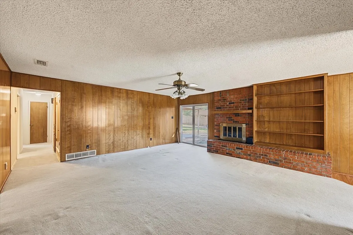 Brick fireplace with built-in shelving, wood paneling, ceiling fan, and sliding glass door