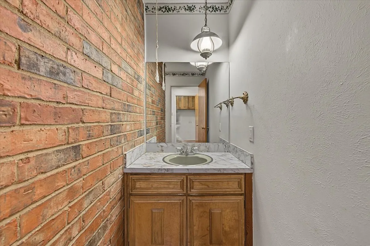 Exposed brick wall, single vanity with wood-finish cabinetry, countertop with a marbled pattern, and a built-in sink