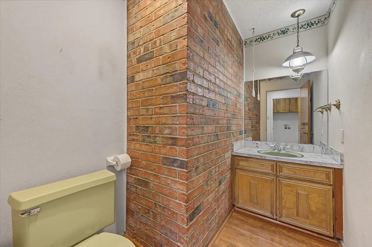 Exposed brick wall detail, wood-finish flooring, vanity with light-colored countertop, built-in vanity cabinetry, and a ceiling-mounted light fixture