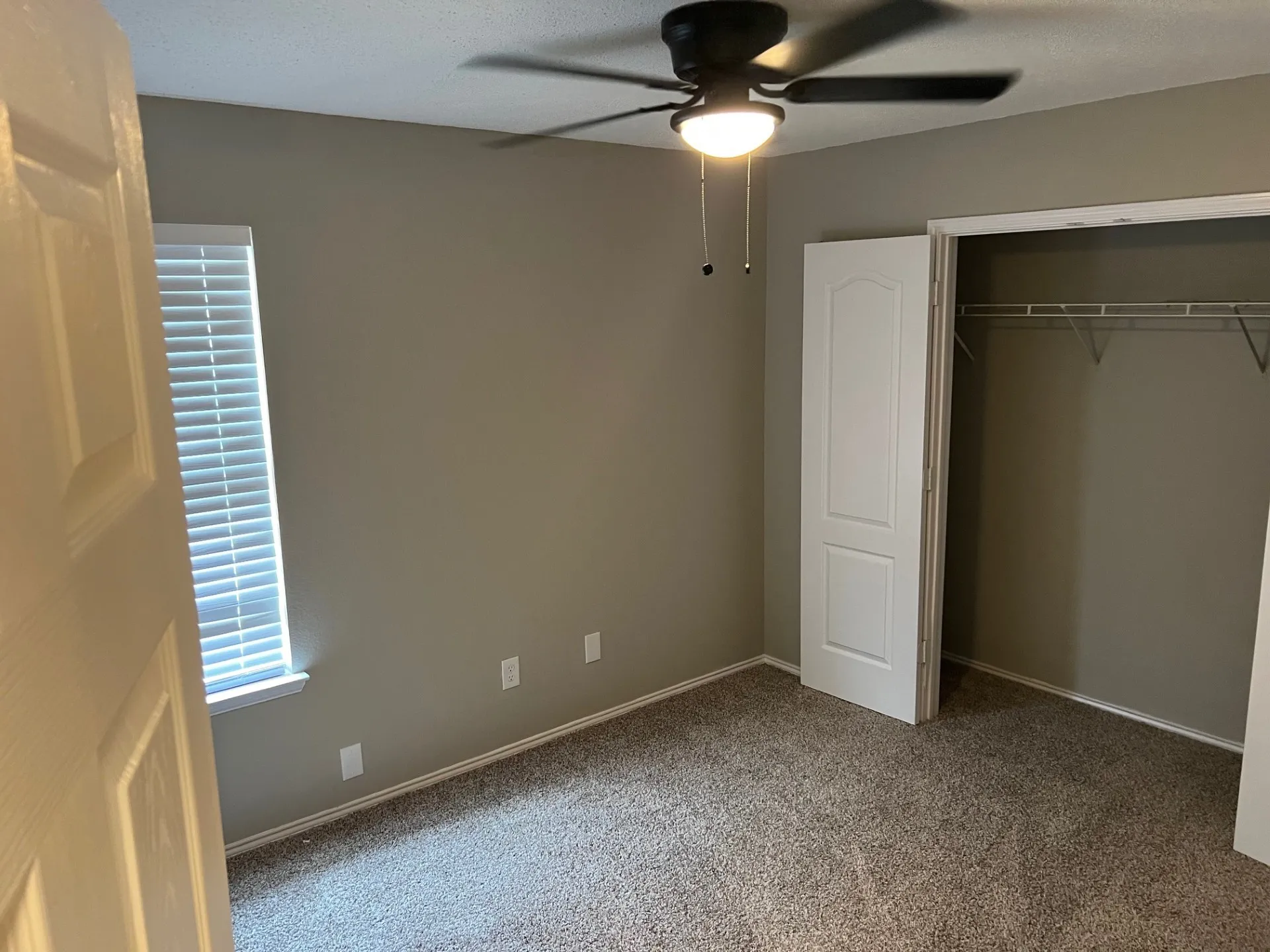 Textured carpeting, neutral wall palette, window with blinds, bi-fold closet doors, and overhead ceiling fan with light