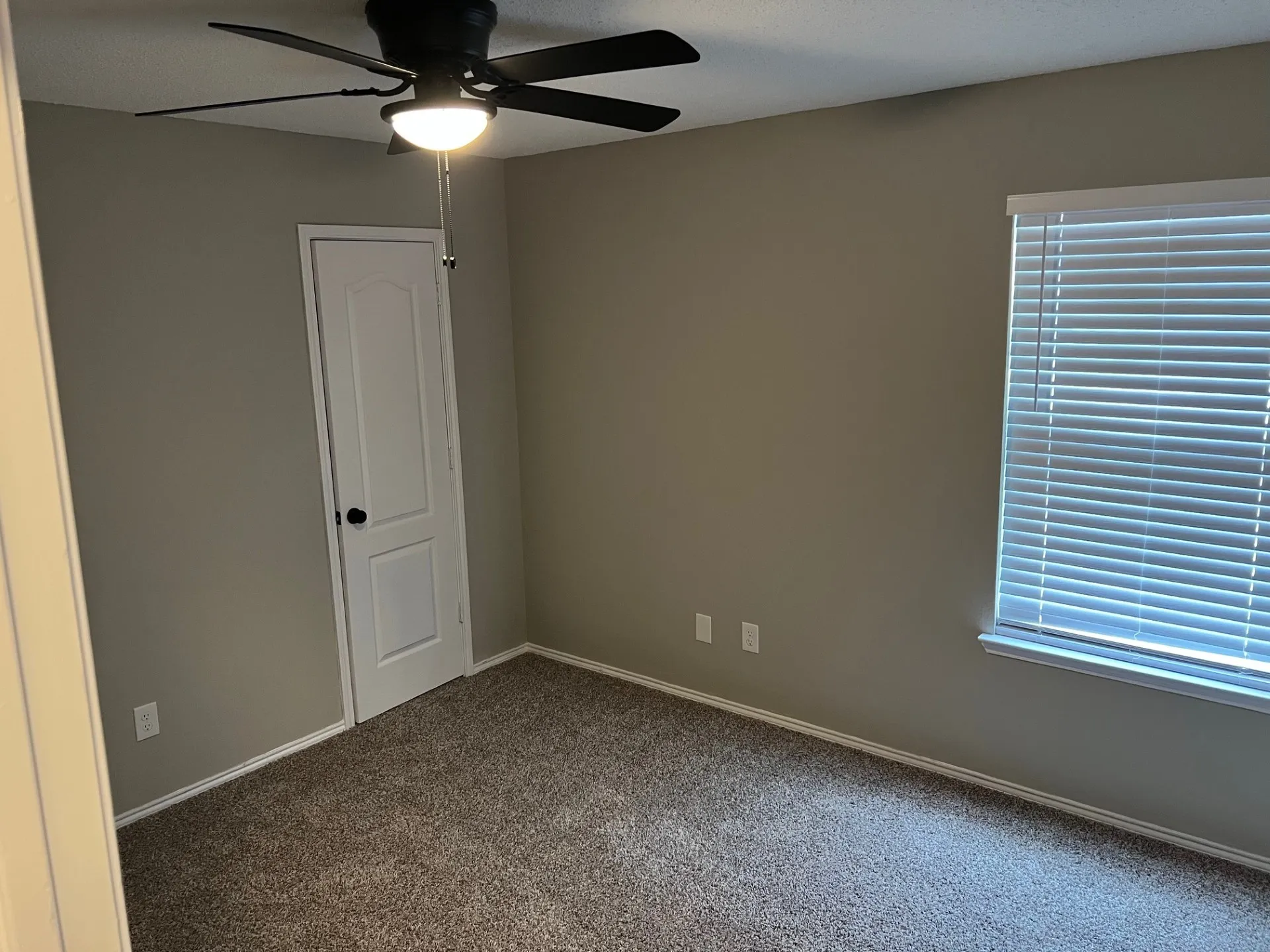 Neutral wall paint, plush carpet flooring, white trim, white door with black hardware, and a ceiling fan with integrated lighting
