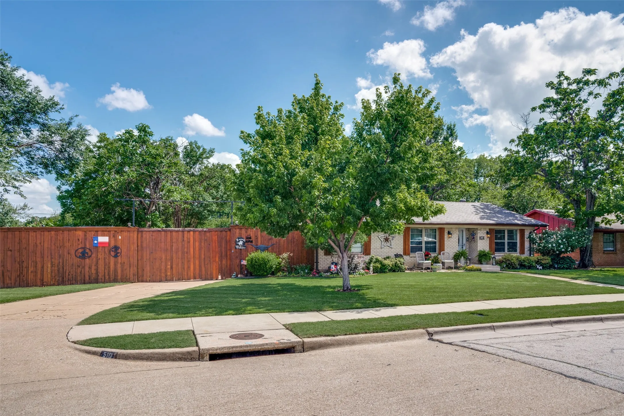 Manicured front yard with mature trees, a paved driveway, and a wood privacy fence
