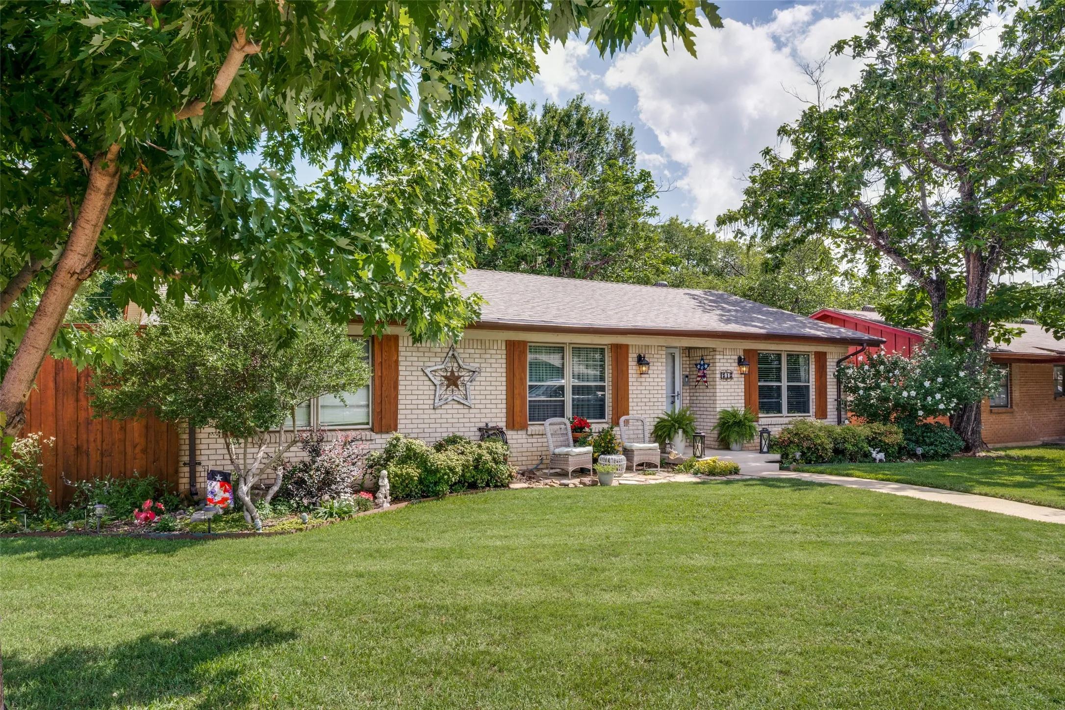 Manicured front lawn with mature trees, brick exterior, front porch, and wood-finish shutters
