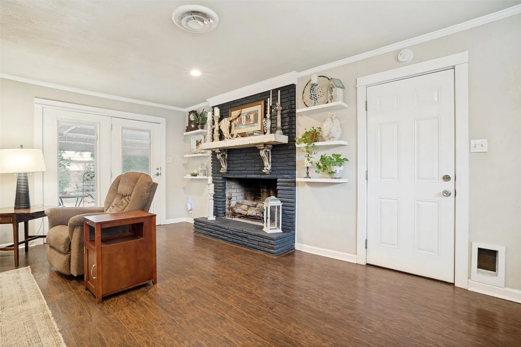 Wood-like flooring, brick fireplace with a mantel, built-in shelving, white paneled door, and double French doors
