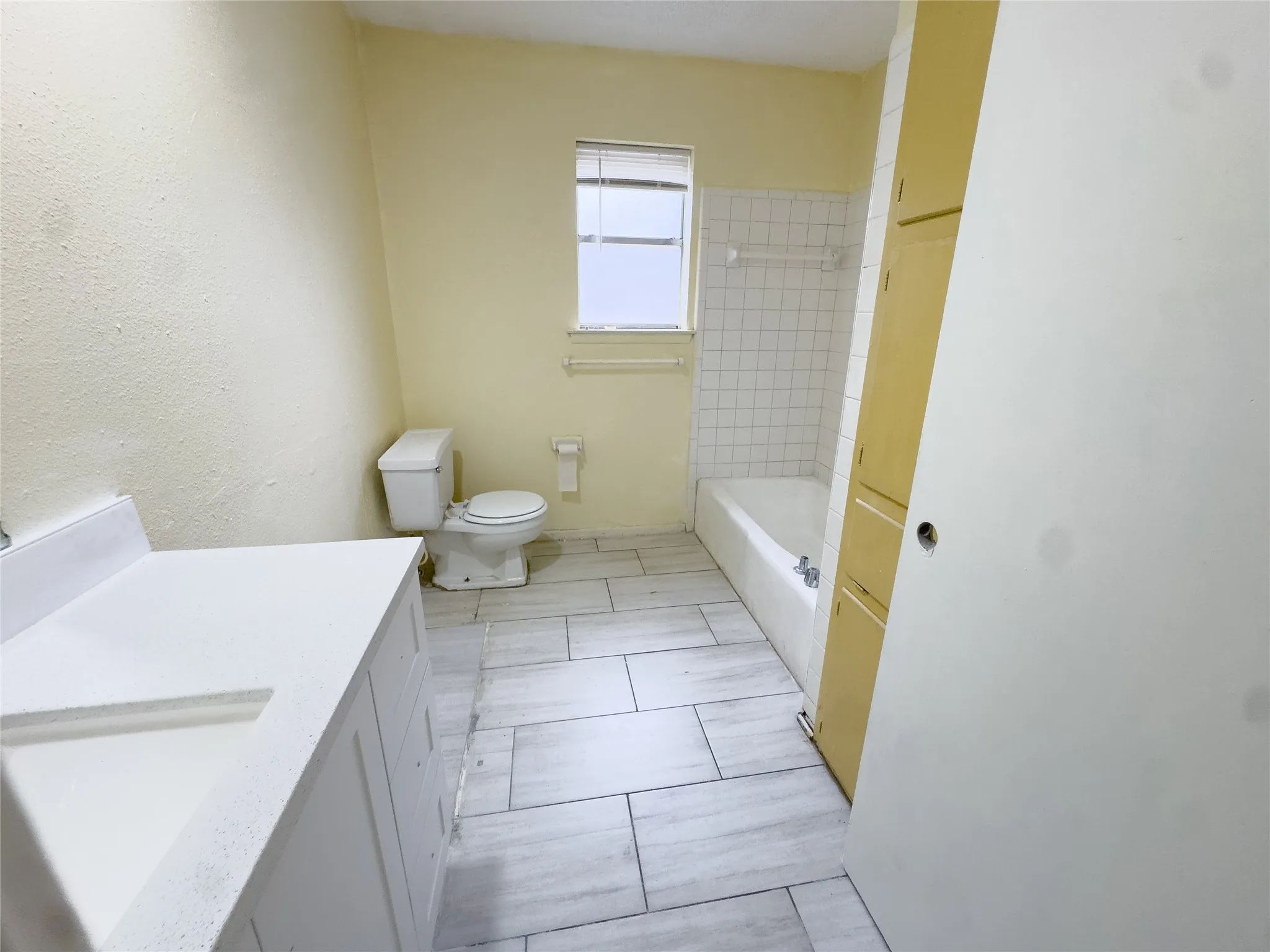 White vanity with an integrated sink, light-toned wood-finish tile flooring, recessed built-in shelving, white bathtub with wall tile surround, and a single window