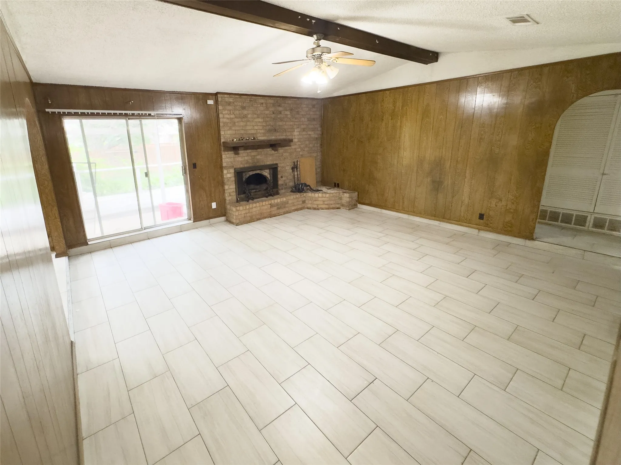 Light-toned tile flooring, brick fireplace with hearth, wood paneling, exposed ceiling beam, and sliding glass doors