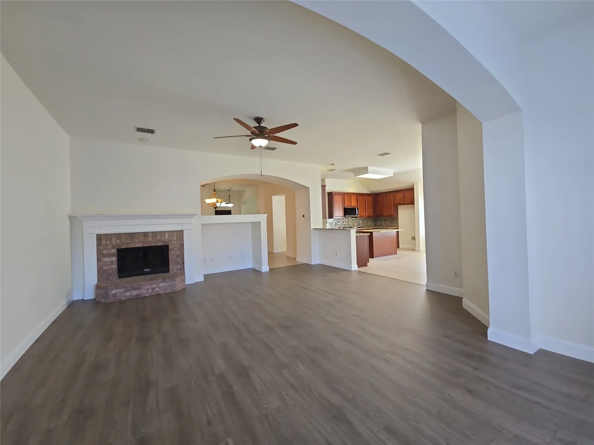 Spacious interior with wood-finish flooring, a brick fireplace with white mantel, a ceiling fan, and arched openings