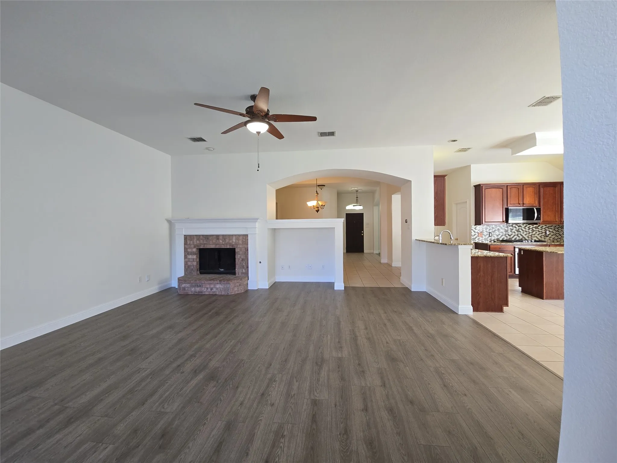 Spacious living area featuring wood-finish flooring, a brick fireplace with a white mantel, and an overhead ceiling fan