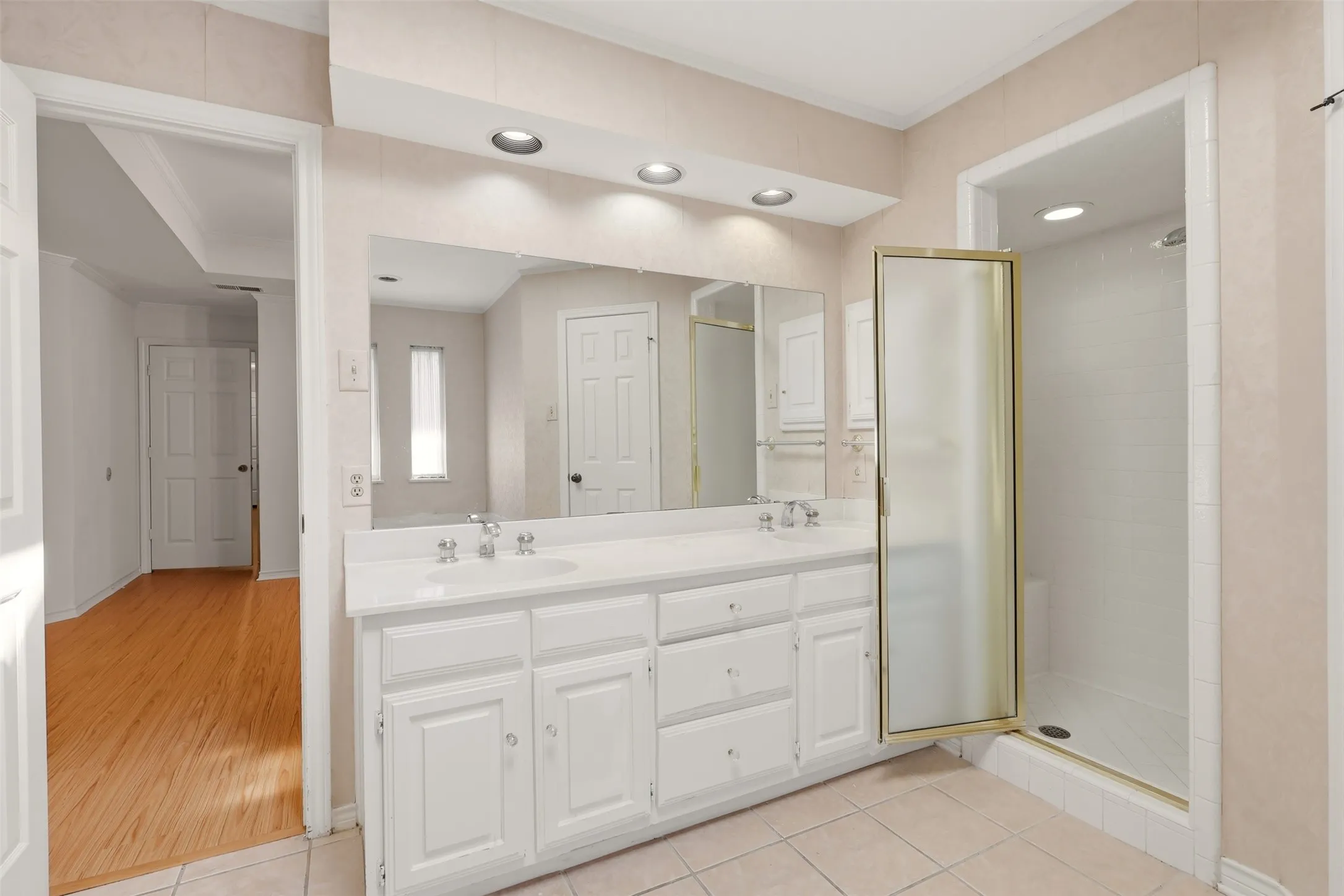 Dual vanity with integrated sinks, recessed lighting, expansive wall mirror, and an enclosed shower with a folding glass door