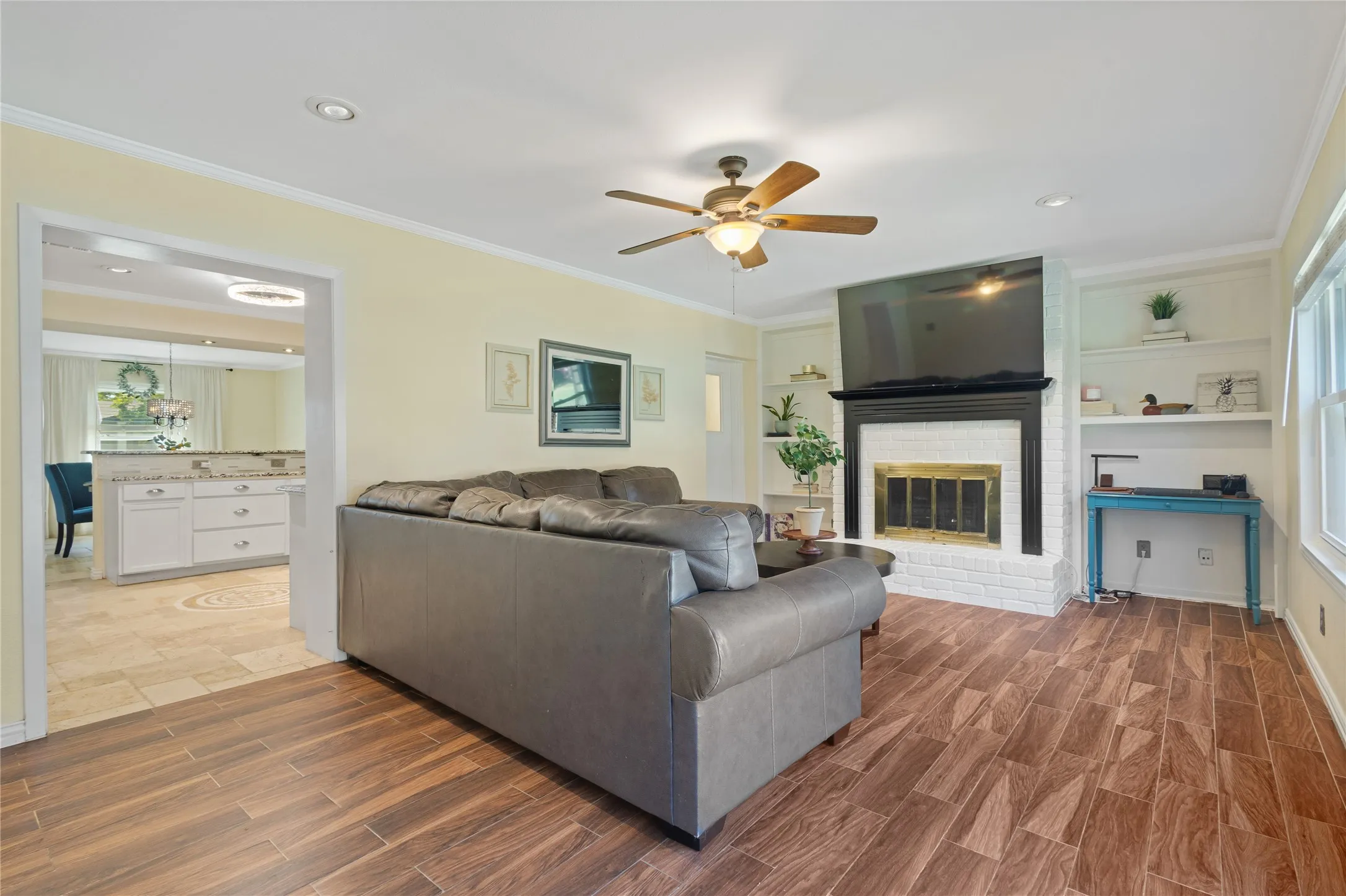 Wood-finish flooring, white brick fireplace with mantel, recessed lighting, ceiling fan, and built-in shelving