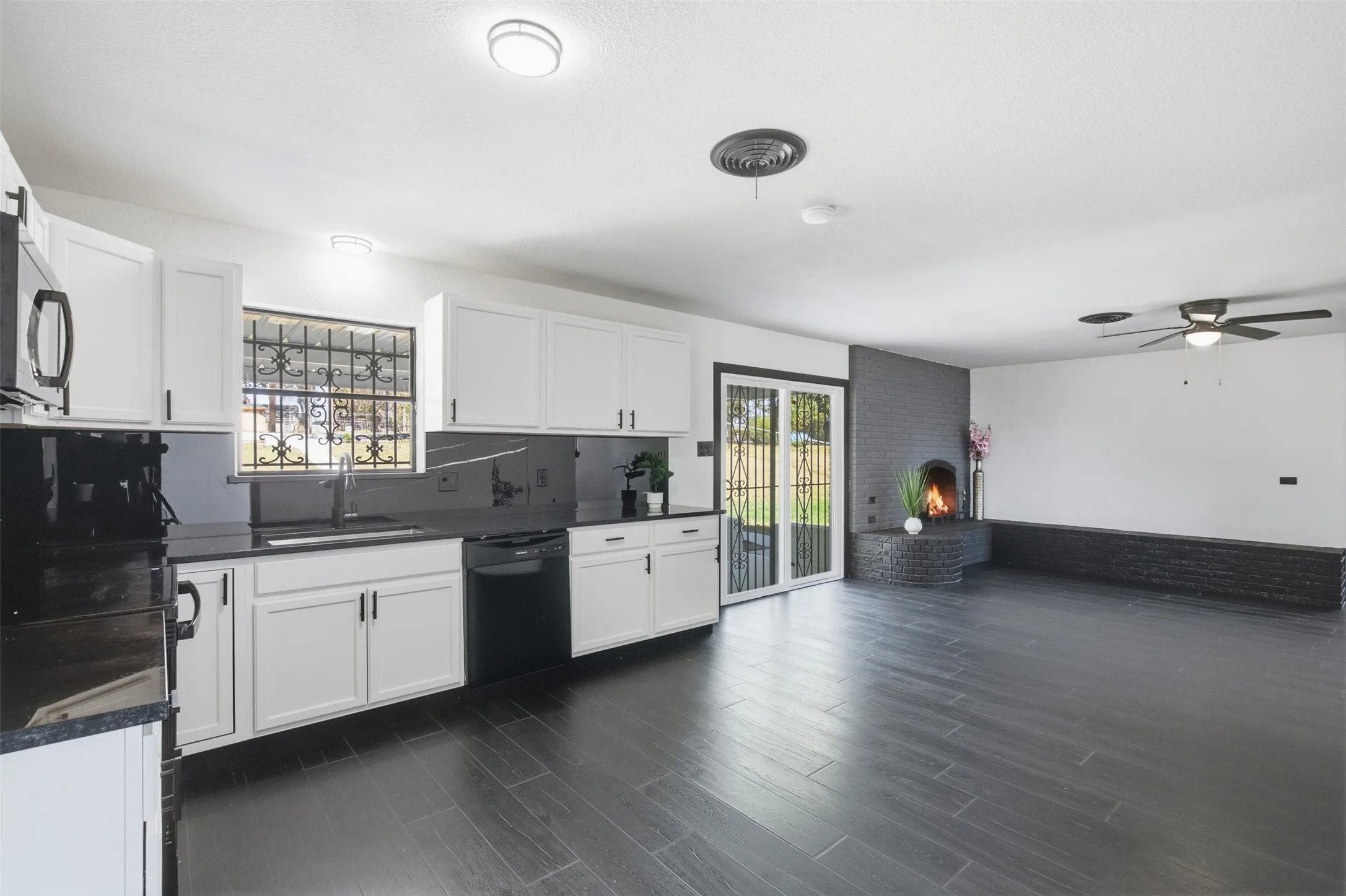 Kitchen and living area featuring dark tile flooring, white cabinetry, dark countertops, a dark brick fireplace, and a ceiling fan