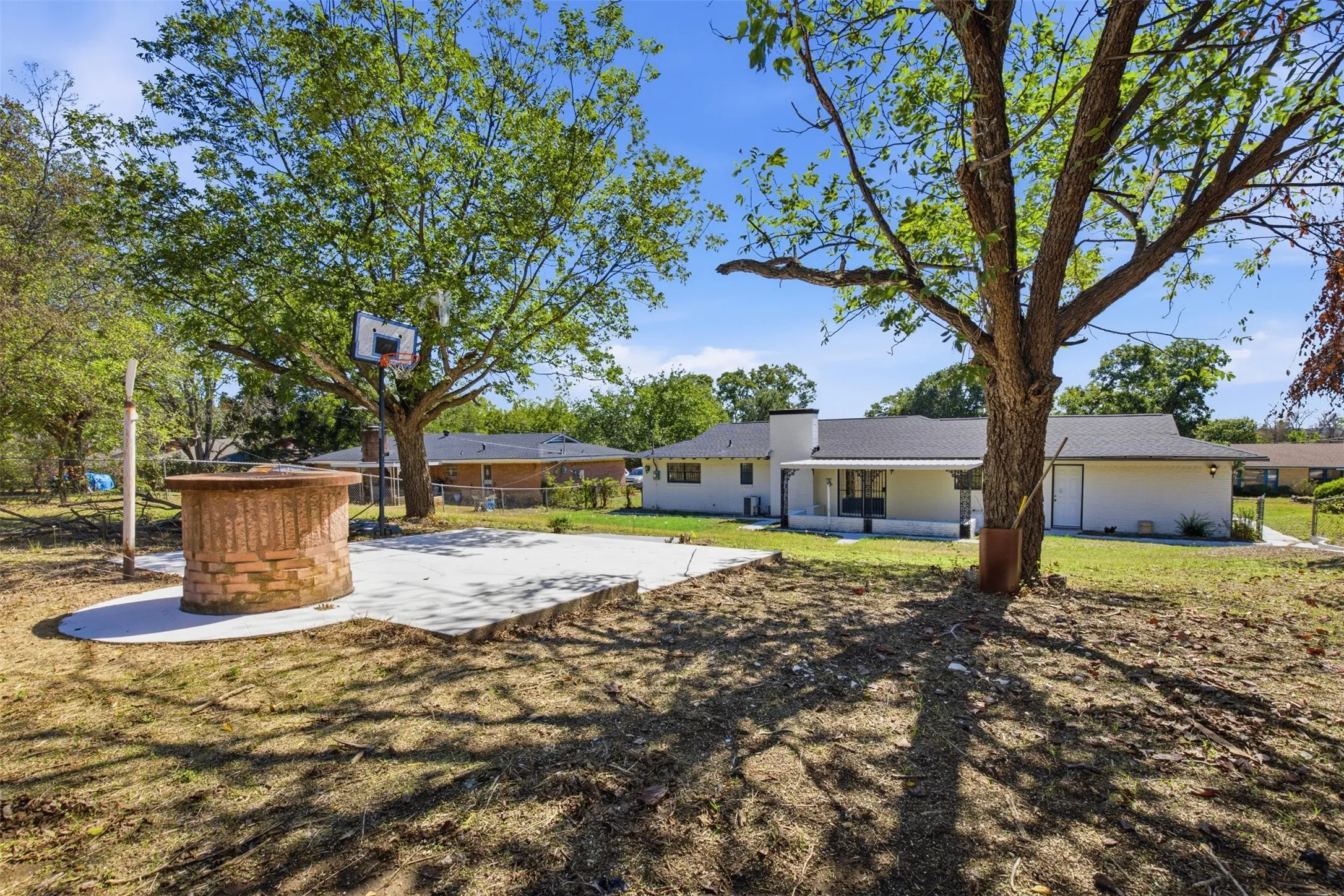 Expansive backyard featuring a concrete patio, a built-in brick fire pit, and mature trees providing natural shade