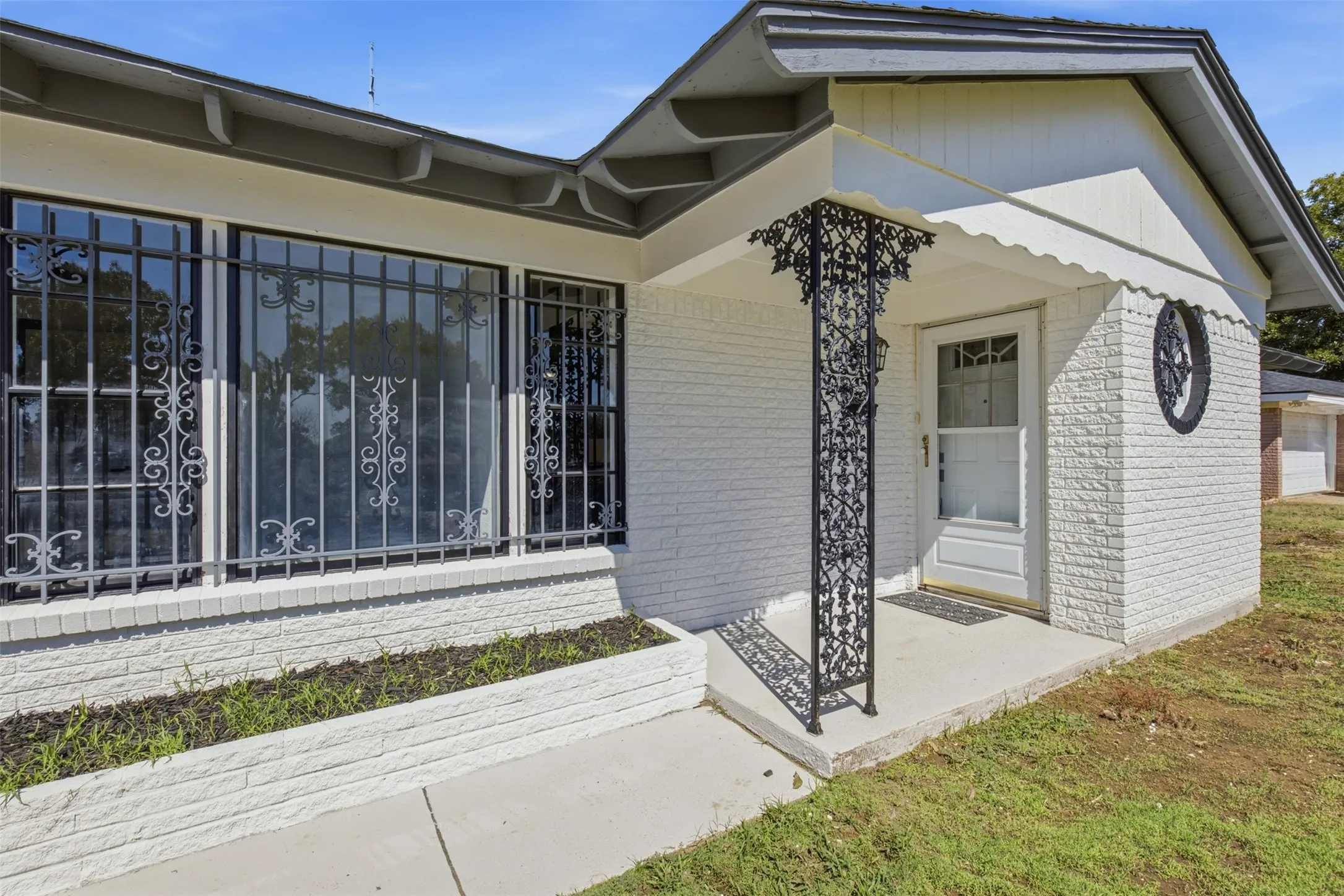 White brick exterior with black ornamental window grilles, a decorative porch column, and a white front entry door with a storm door