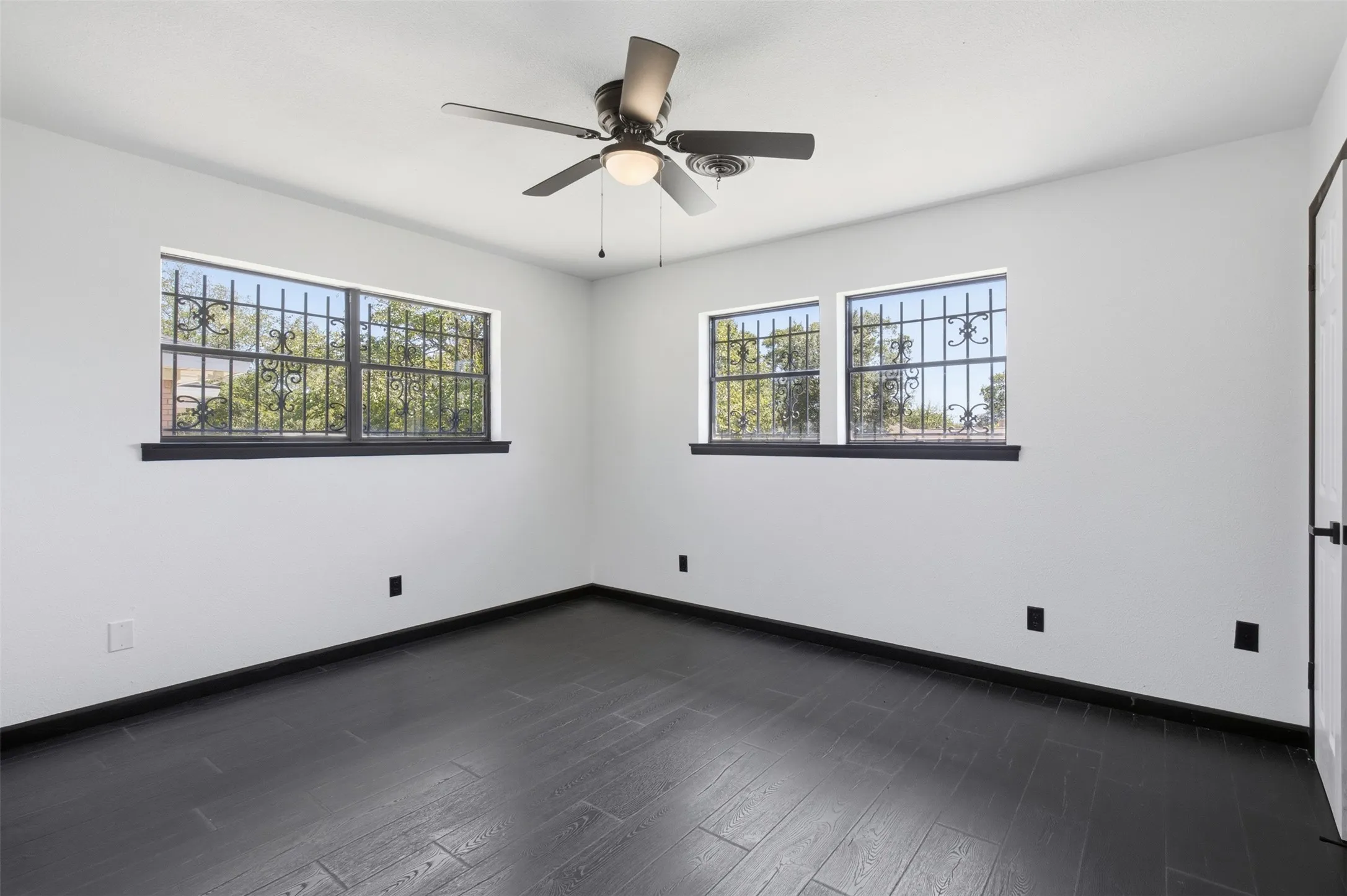 Wood-finish flooring, two windows with decorative ironwork, and a ceiling fan with integrated lighting
