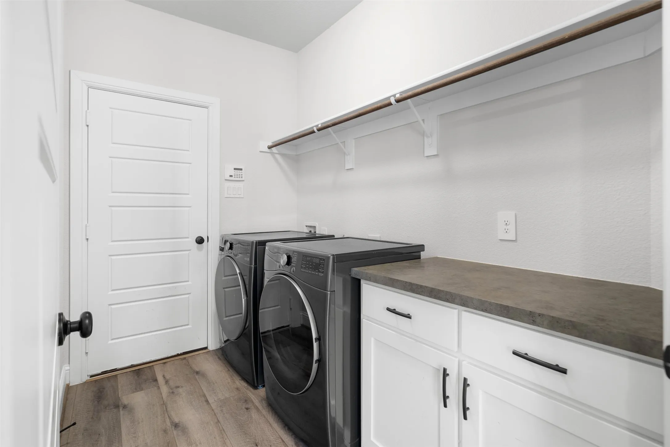 Laundry area featuring wood-finish flooring, white cabinetry with black hardware, a grey countertop, and an overhead clothes rod
