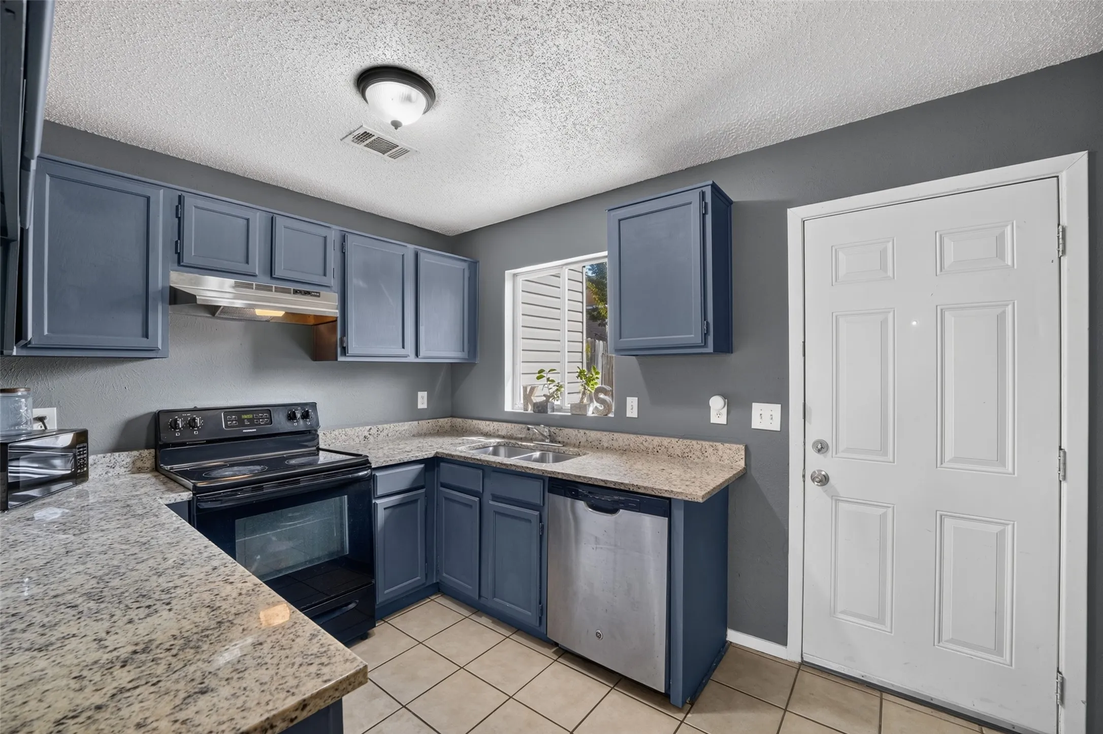 Kitchen featuring granite-style countertops, dark-toned cabinetry, stainless steel sink, tile flooring, and a white paneled door