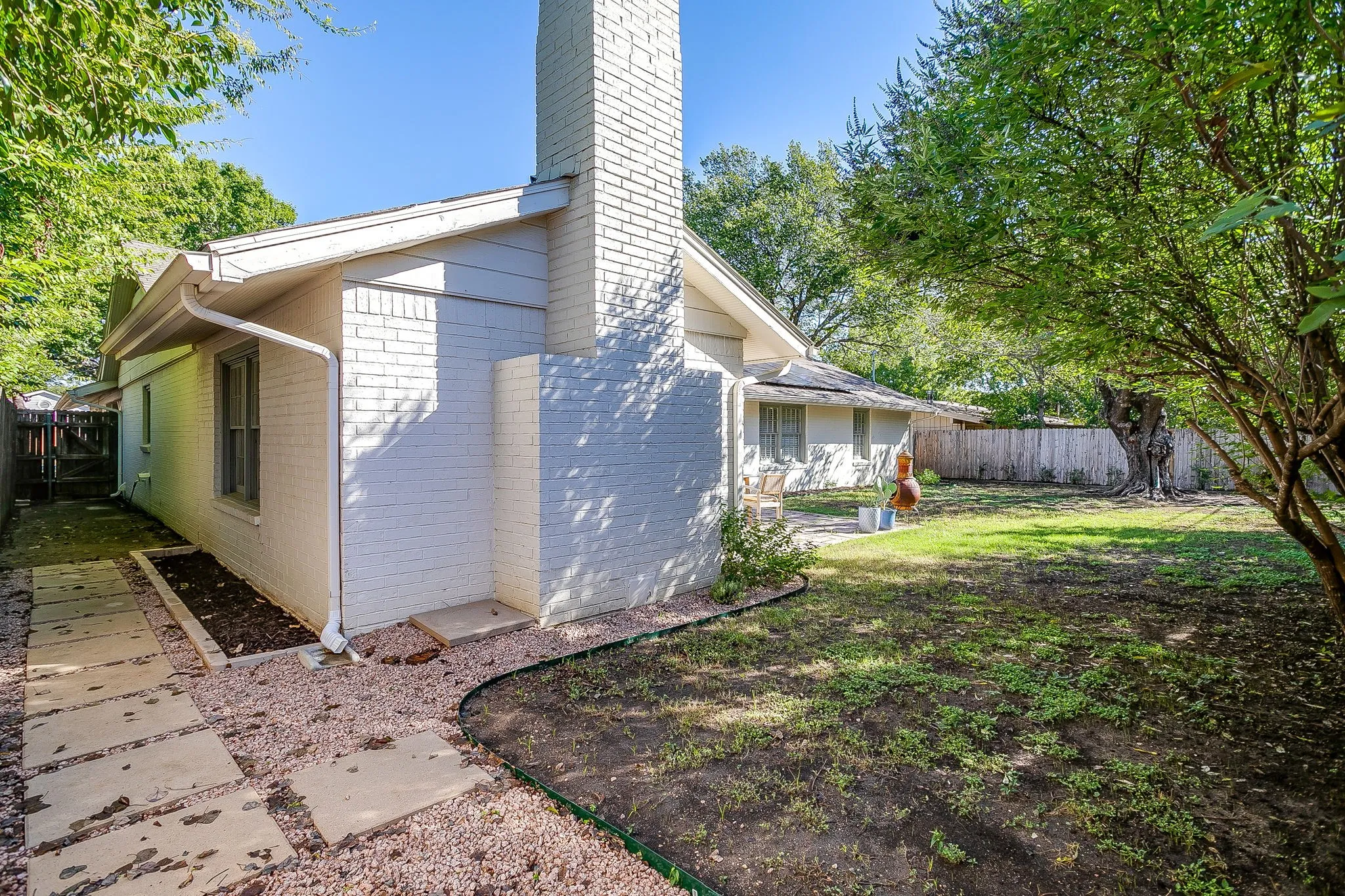 White brick exterior with a prominent brick chimney, accompanied by a spacious lawn area, partial privacy fencing, and mature trees