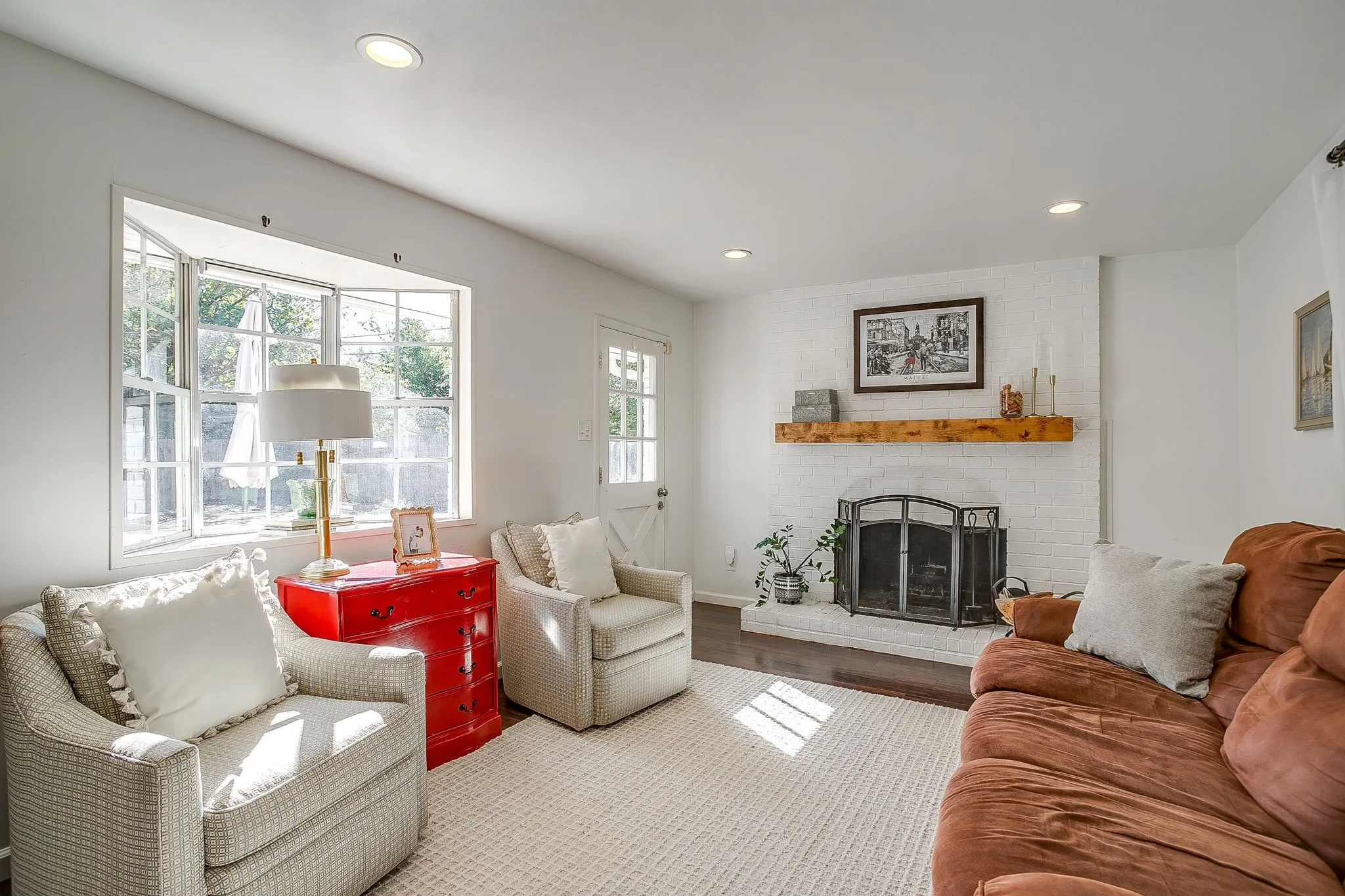 Bright, sun-filled living room featuring multiple large windows with natural light, neutral wall finishes, recessed ceiling lighting, wood-finish flooring with neutral area rug, and a fireplace with dark surround and built-in cabinetry flanking the hearth