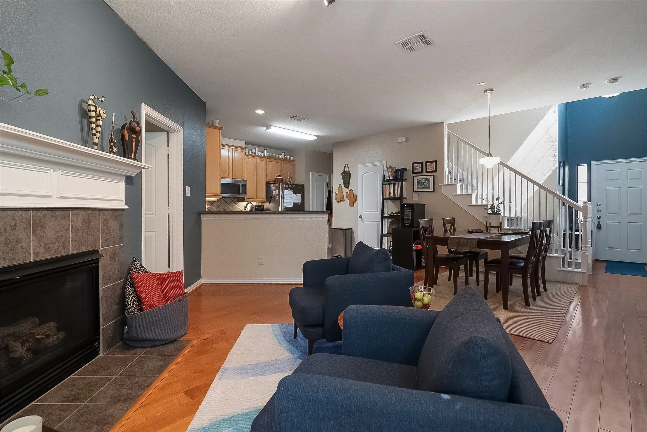 Fireplace with tile surround, wood-finish flooring, kitchen with wood cabinetry, staircase with white spindles, and an entryway with a light-colored door and sidelights