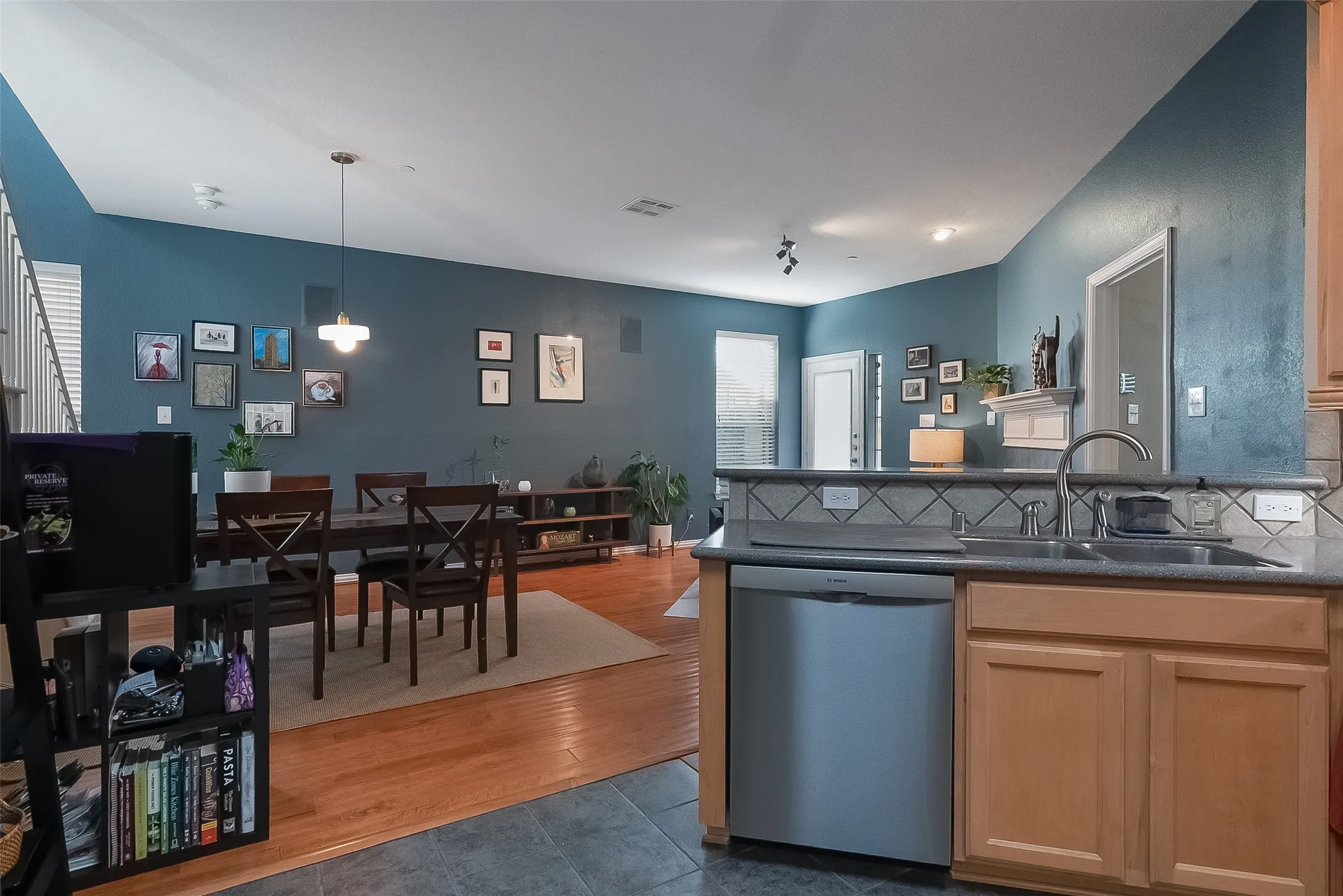 Open-concept common area featuring wood-finish flooring, a kitchen with light wood cabinetry, stainless steel dishwasher, and a double basin sink