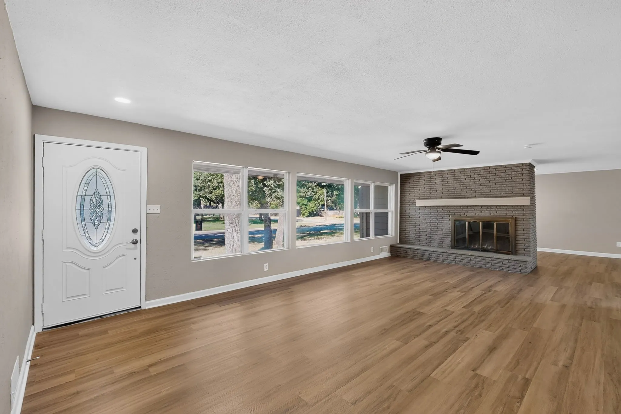 Spacious living area featuring wood-finish flooring, a brick fireplace with a mantle, a ceiling fan with integrated lighting, and multiple windows