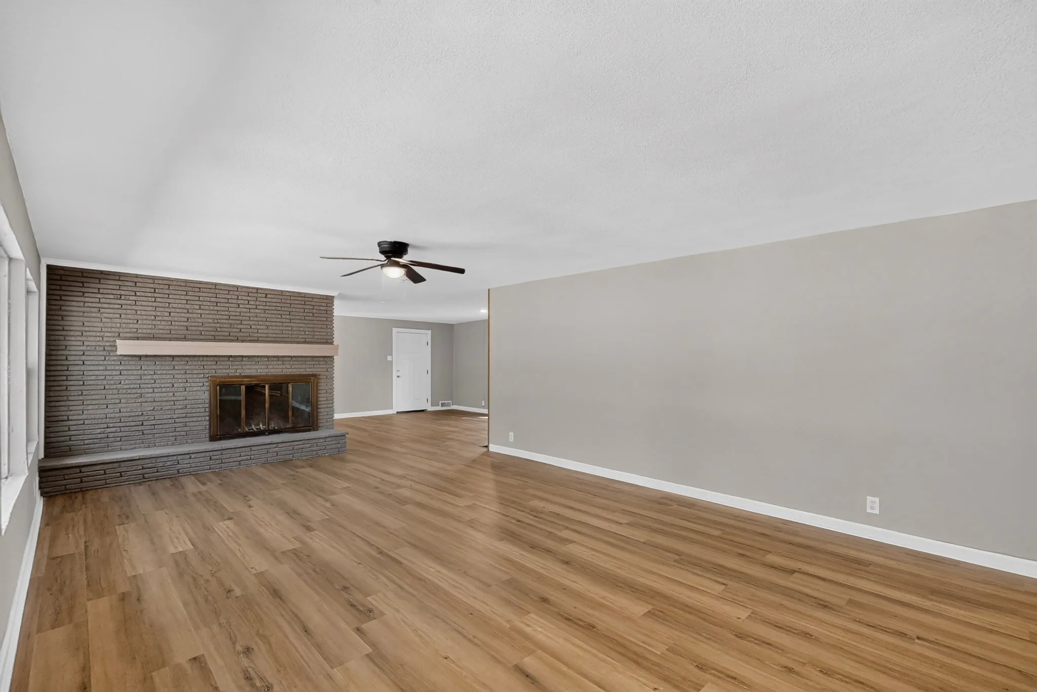 Expansive room with wood-finish flooring, a brick fireplace with a wood mantle, and a ceiling fan