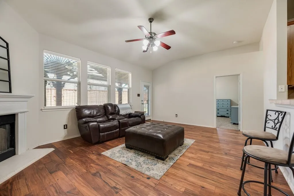 Vaulted ceiling living area featuring wood-finish flooring, a white mantel, and a multi-blade ceiling fan