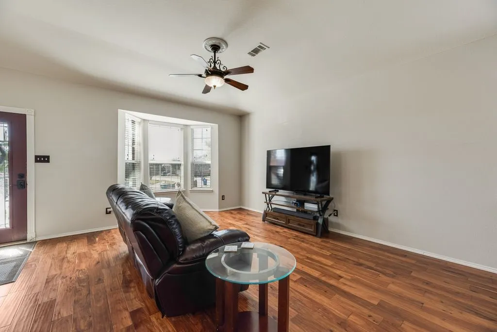 Wood-finish flooring throughout, bay window with blinds, ceiling fan, and a red-finish entry door with decorative glass