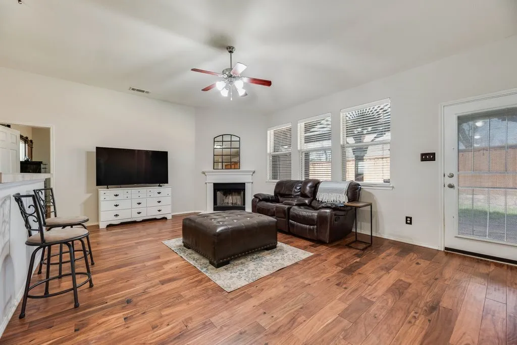 Wood-finish flooring throughout, vaulted ceiling, ceiling fan, white paneled fireplace, and multiple windows with blinds