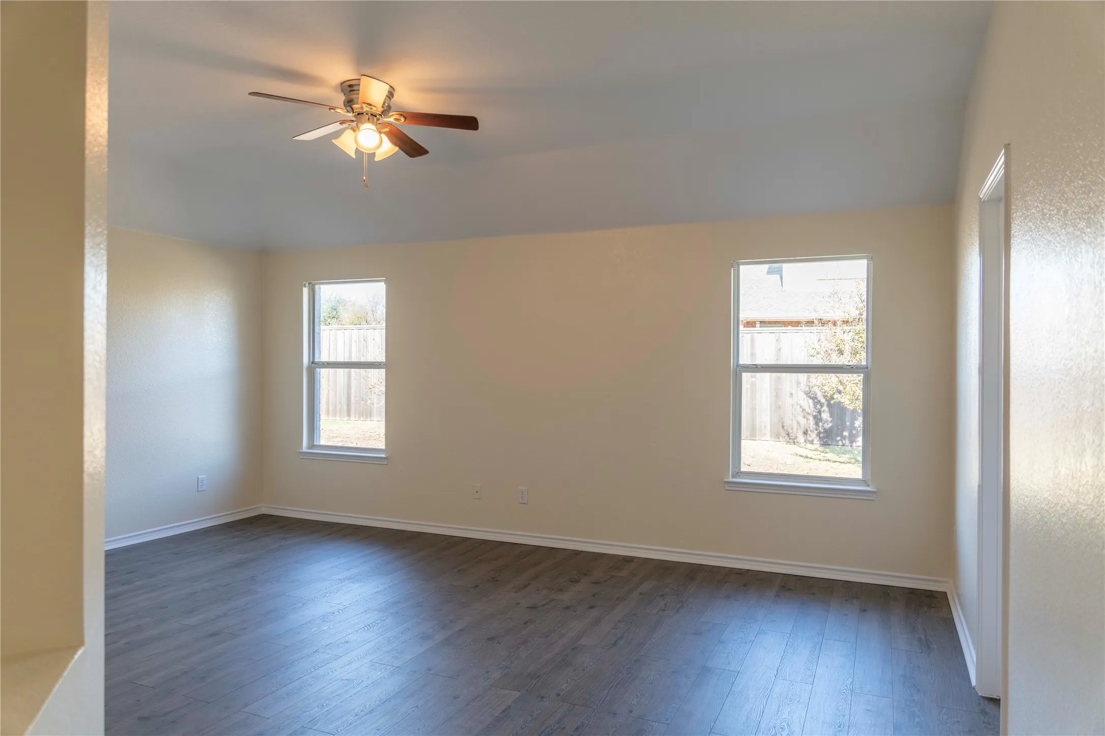 Spacious room featuring wood-finish flooring, two large windows, a ceiling fan with integrated lighting, and neutral wall tones