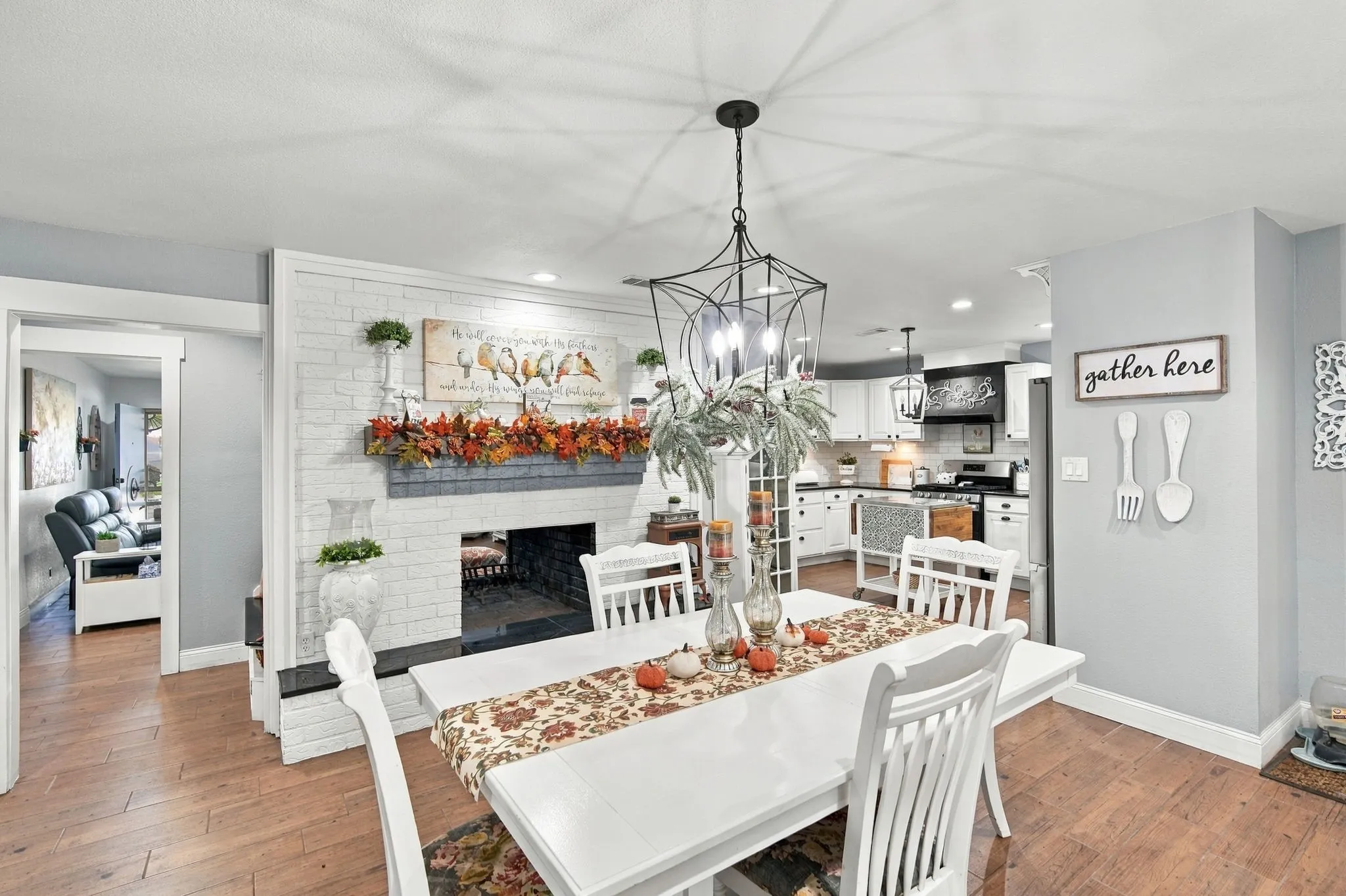 Wood-finish flooring, white brick doujble sided  fireplace with dark mantel, open concept kitchen, white cabinetry, and pendant lighting