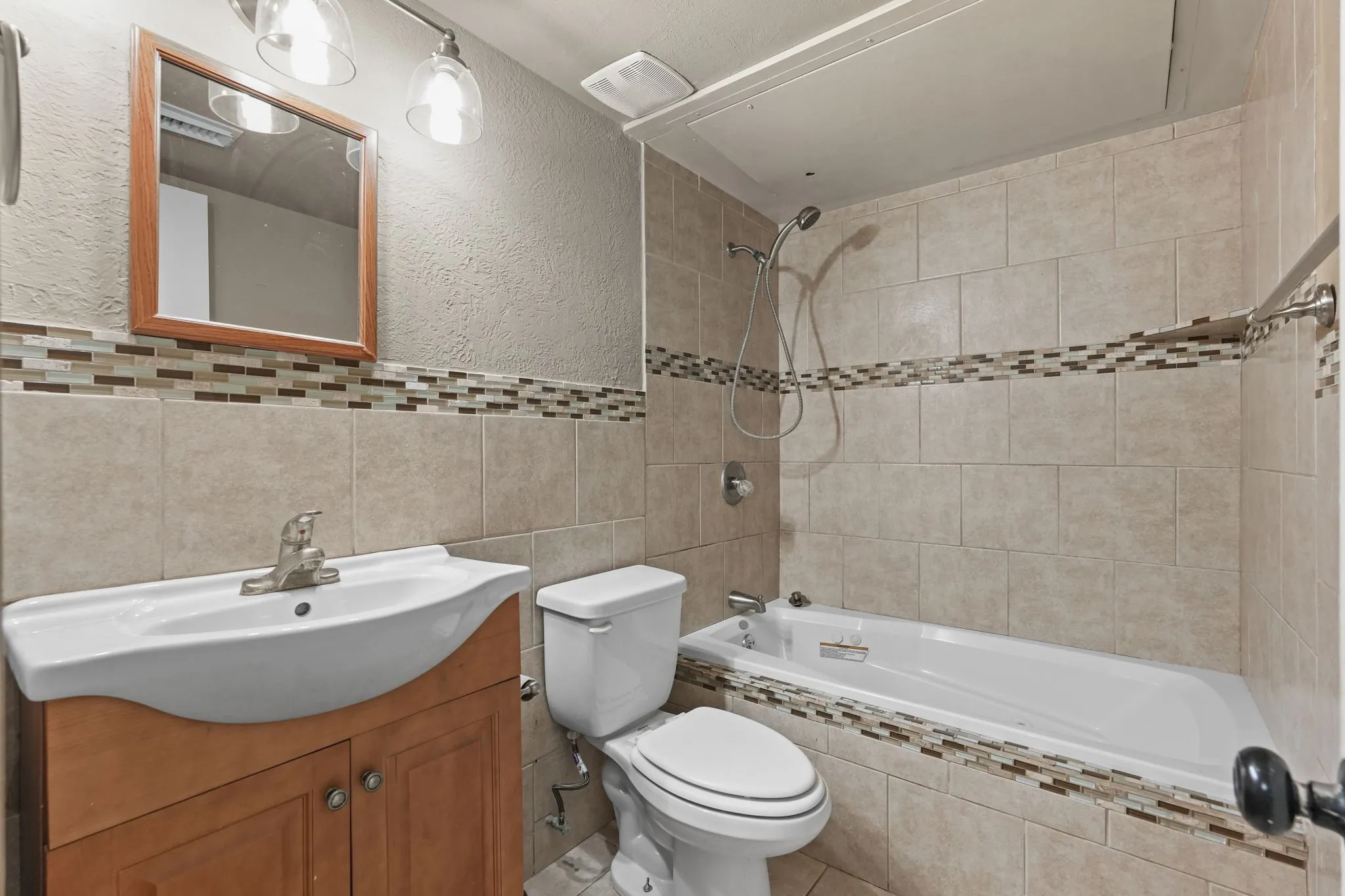 Spa-inspired bathroom featuring a wood-finish vanity with dual-basin sink, ceramic tile walls with decorative tile accent band, recessed ceiling lighting, an alcove bathtub with shower enclosure and rainfall showerhead, and white ceramic toilet