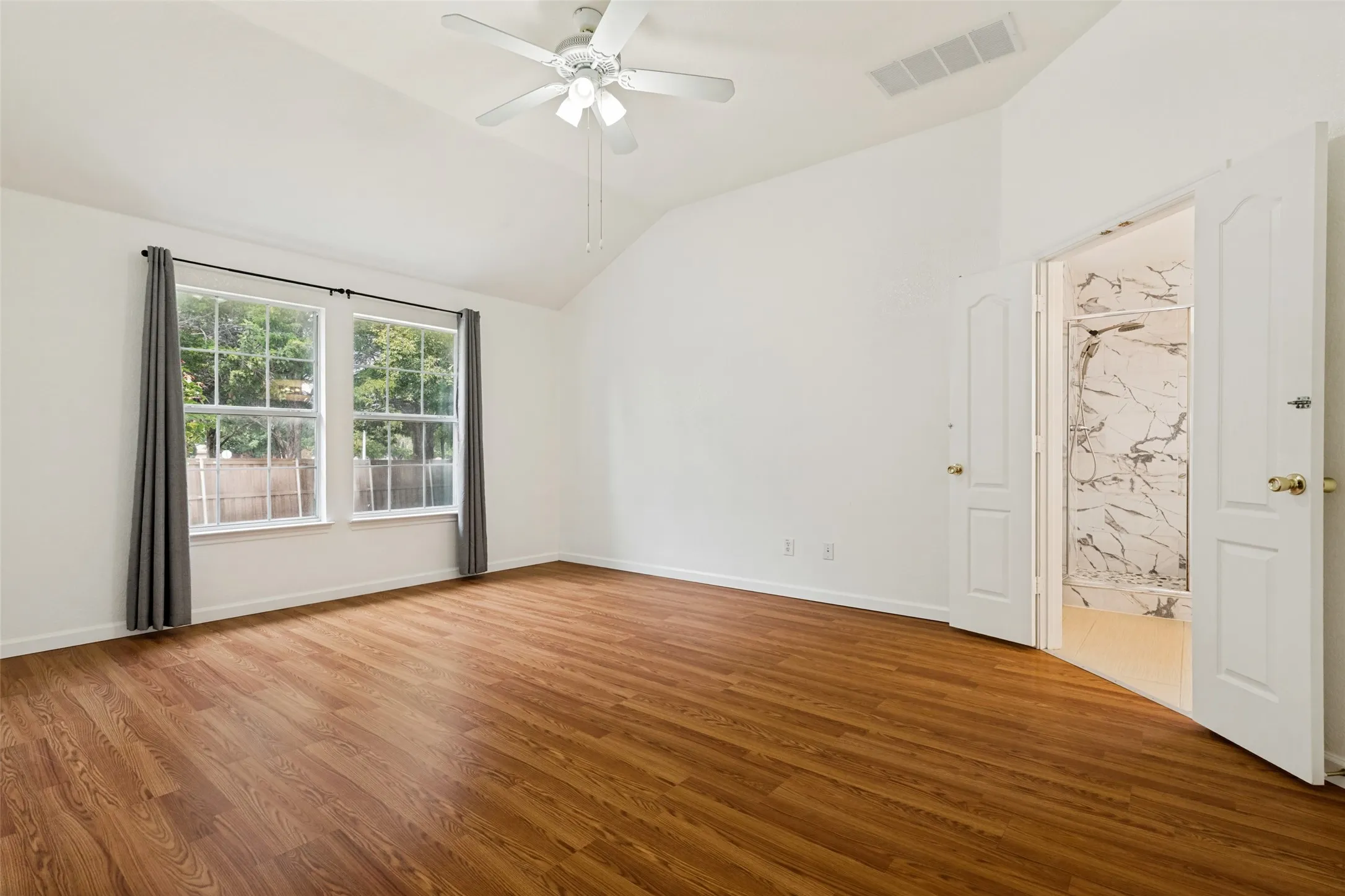 Vaulted ceiling with a ceiling fan, dual windows, wood-finish flooring, and an open doorway to a shower with white and gray veined tile